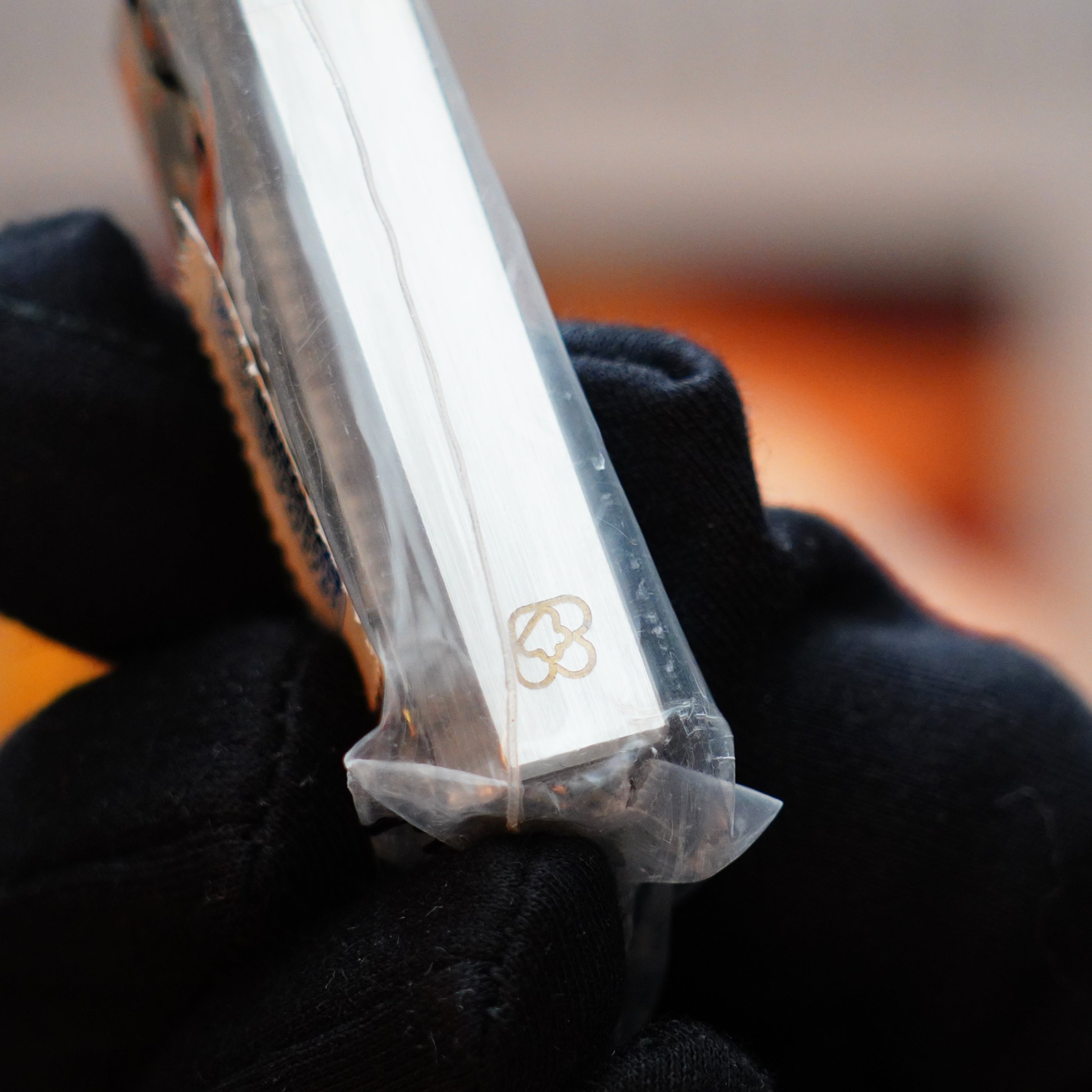 Close-up of a gloved hand holding a rare Mini size silver metallic object, the Vintage 1982 Cartier Santos Pentagon Palladium Sealed with solid 18k Gold Real Screw Initial Lighter, featuring an embossed logo resembling a stylized heart on one side.