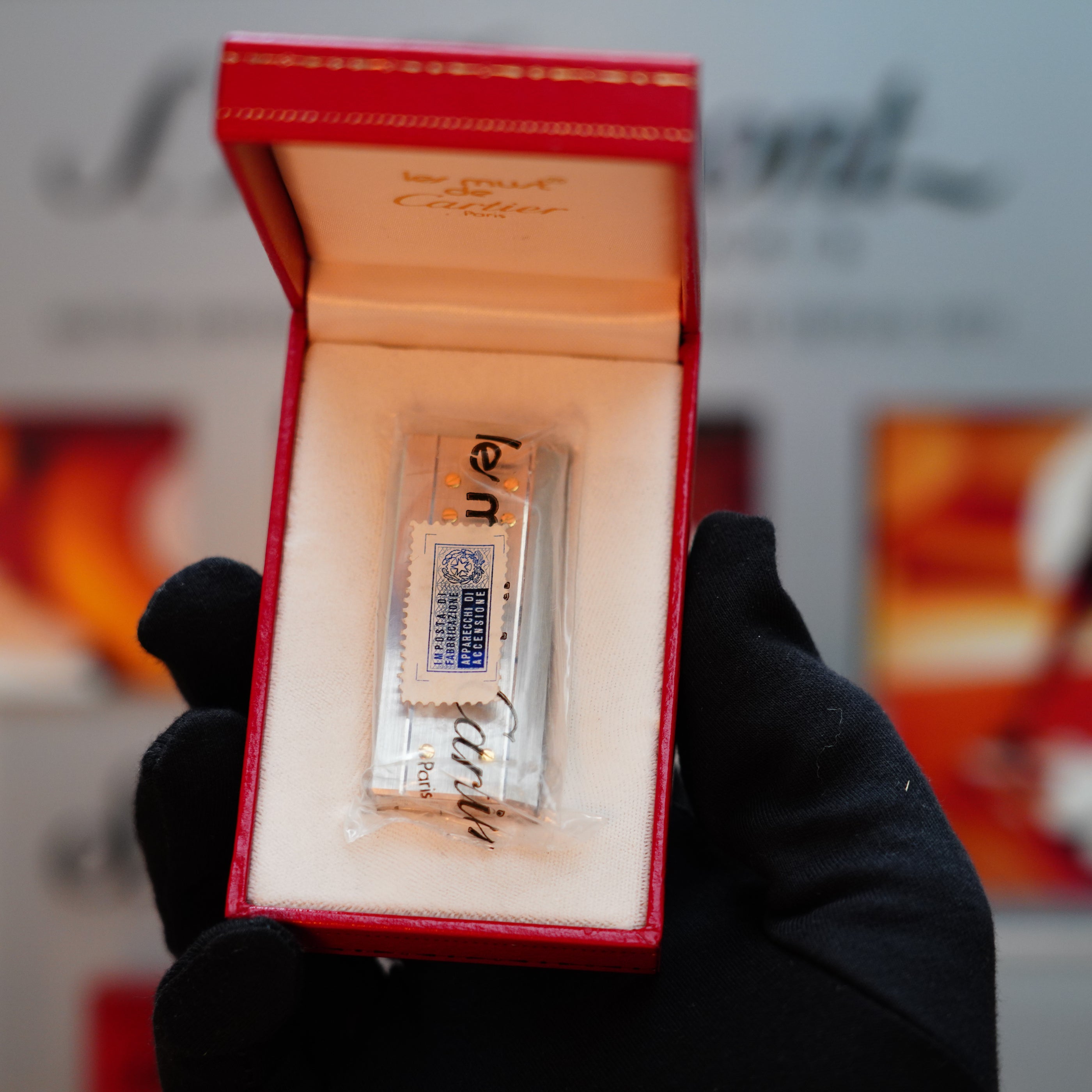 A gloved hand holds an open red jewelry box, revealing a rare, mini-sized, sealed object branded by Cartier. Inside lies a vintage 1982 Cartier Santos Pentagon Palladium lighter, embellished with solid 18k gold screws, encapsulating timeless elegance in its compact design.