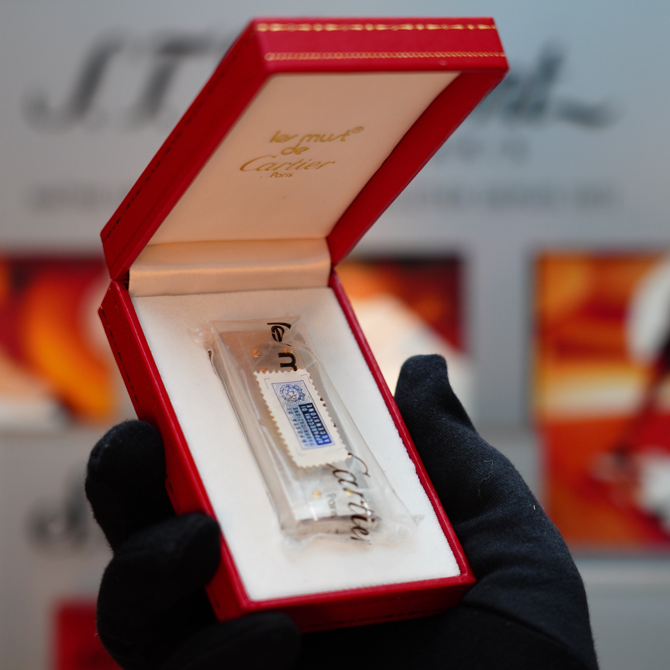 A gloved hand presents a red Cartier box that exudes the luxurious elegance of a Vintage 1982 Cartier Santos Pentagon, sealed with solid 18k gold real screws. Inside, you'll find a rare mini-size item—a clear glass vial with a spray label that epitomizes sophistication.