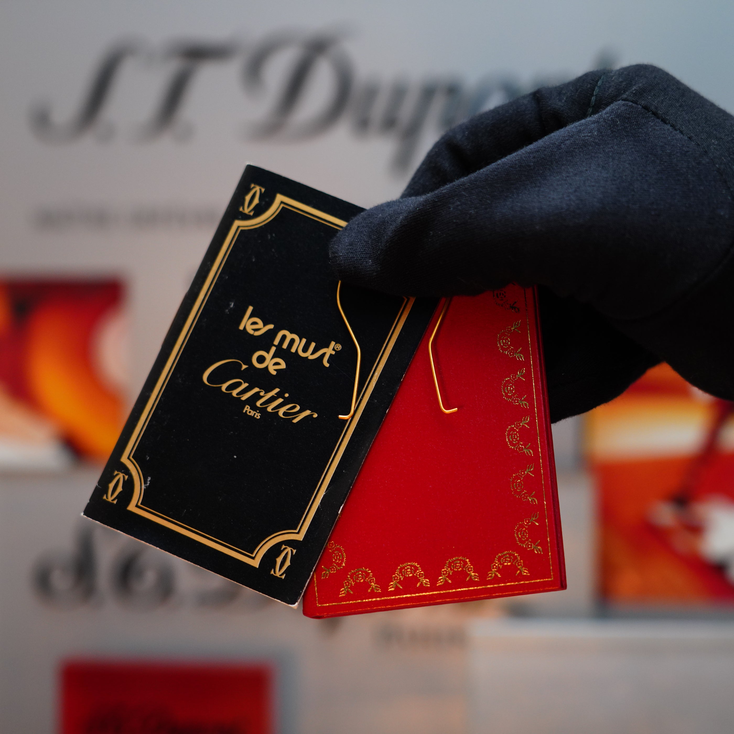 A gloved hand delicately holds two luxury product tags: one black with "le must de Cartier" text and the other red with gold detailing, reminiscent of a rare, vintage 1982 Cartier Santos Pentagon Palladium lighter featuring solid 18k gold screws in a mini size.