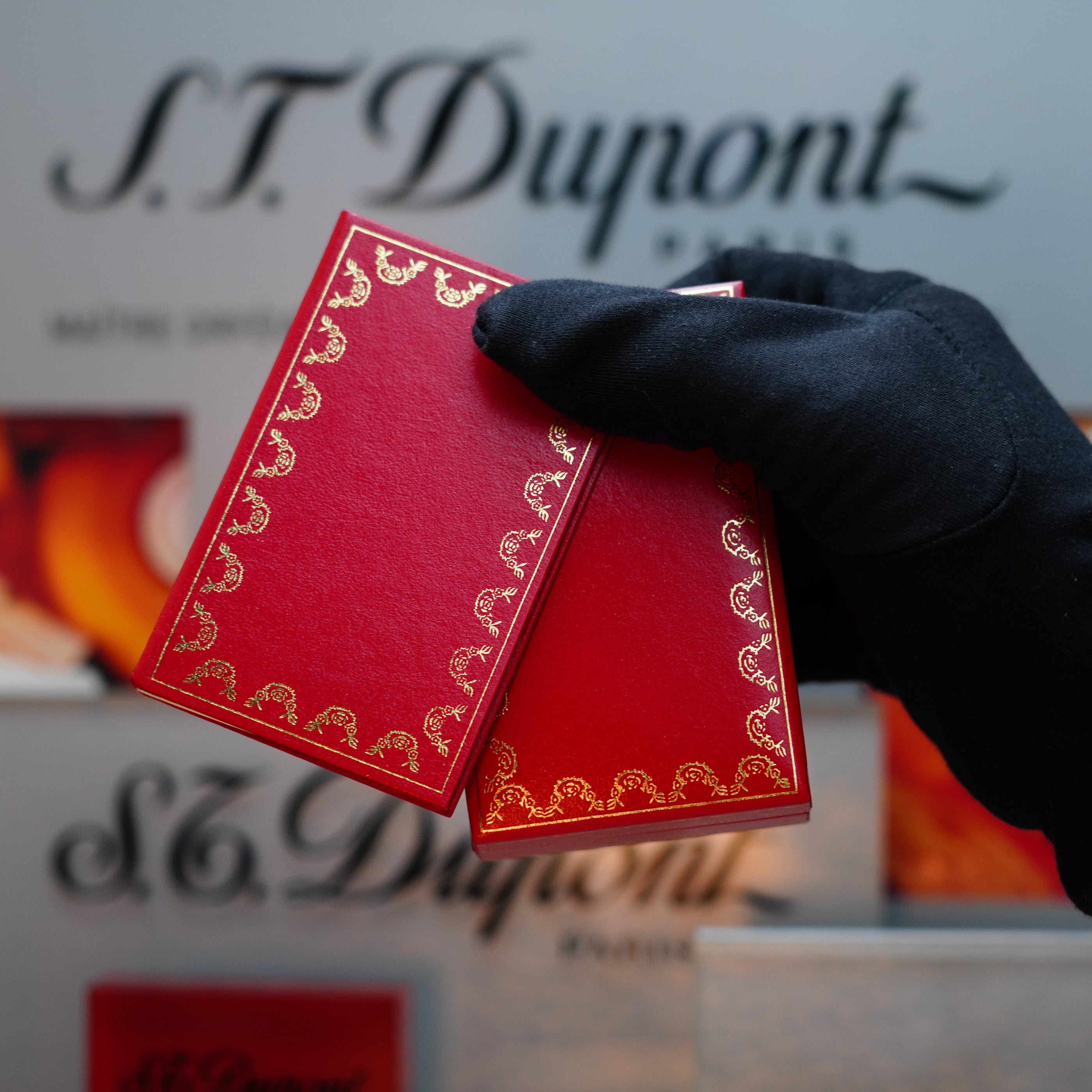 A gloved hand holds two rare mini-size red cards with gold designs, in front of a blurred display featuring the Vintage 1982 Cartier Santos Pentagon Palladium Sealed lighter with solid 18k Gold Real Screw.