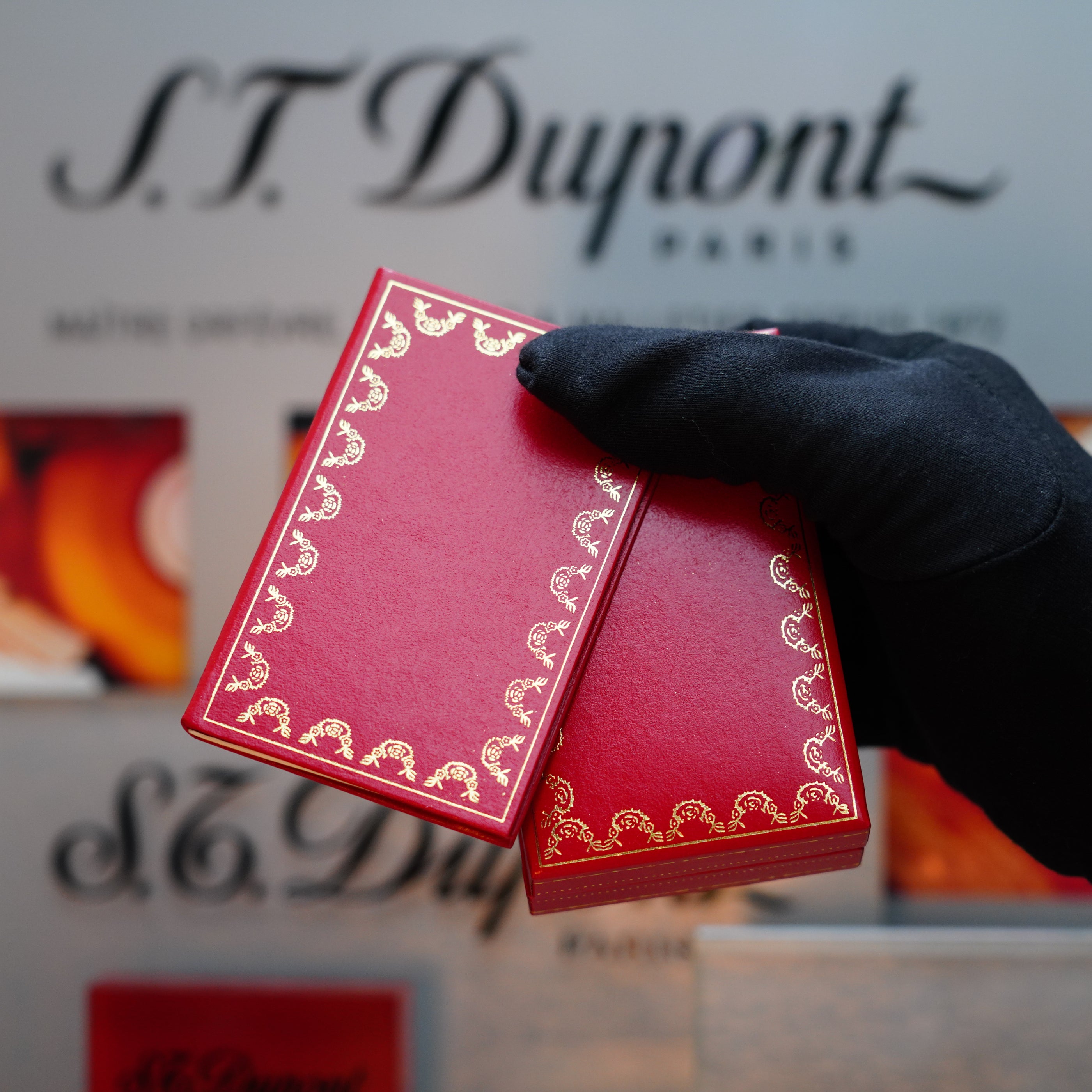 A gloved hand holds two ornate red boxes with gold detailing, reminiscent of a vintage 1982 Cartier Santos Pentagon Palladium Sealed with solid 18k Gold Real Screw, Rare Mini size lighter with an Initial "L", displayed in front of a blurred Cartier sign.