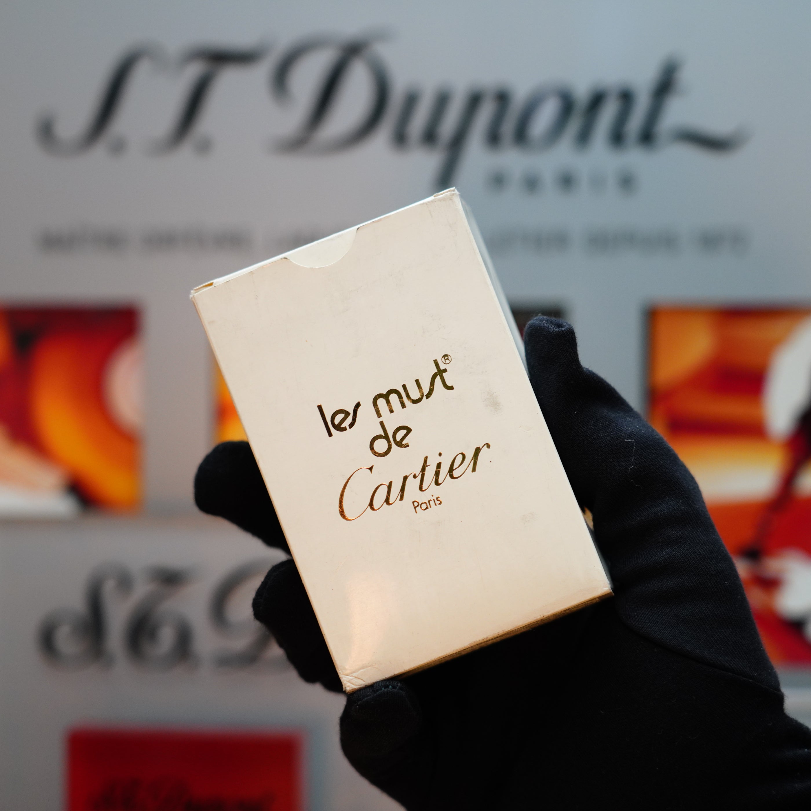 A gloved hand holds a "Le Must de Cartier" box, with the blurred background of S.T. Dupont text hinting at the vintage 1982 Cartier Santos Pentagon Palladium lighter, crafted in a rare mini size with solid 18k gold real screws and initial detailing.