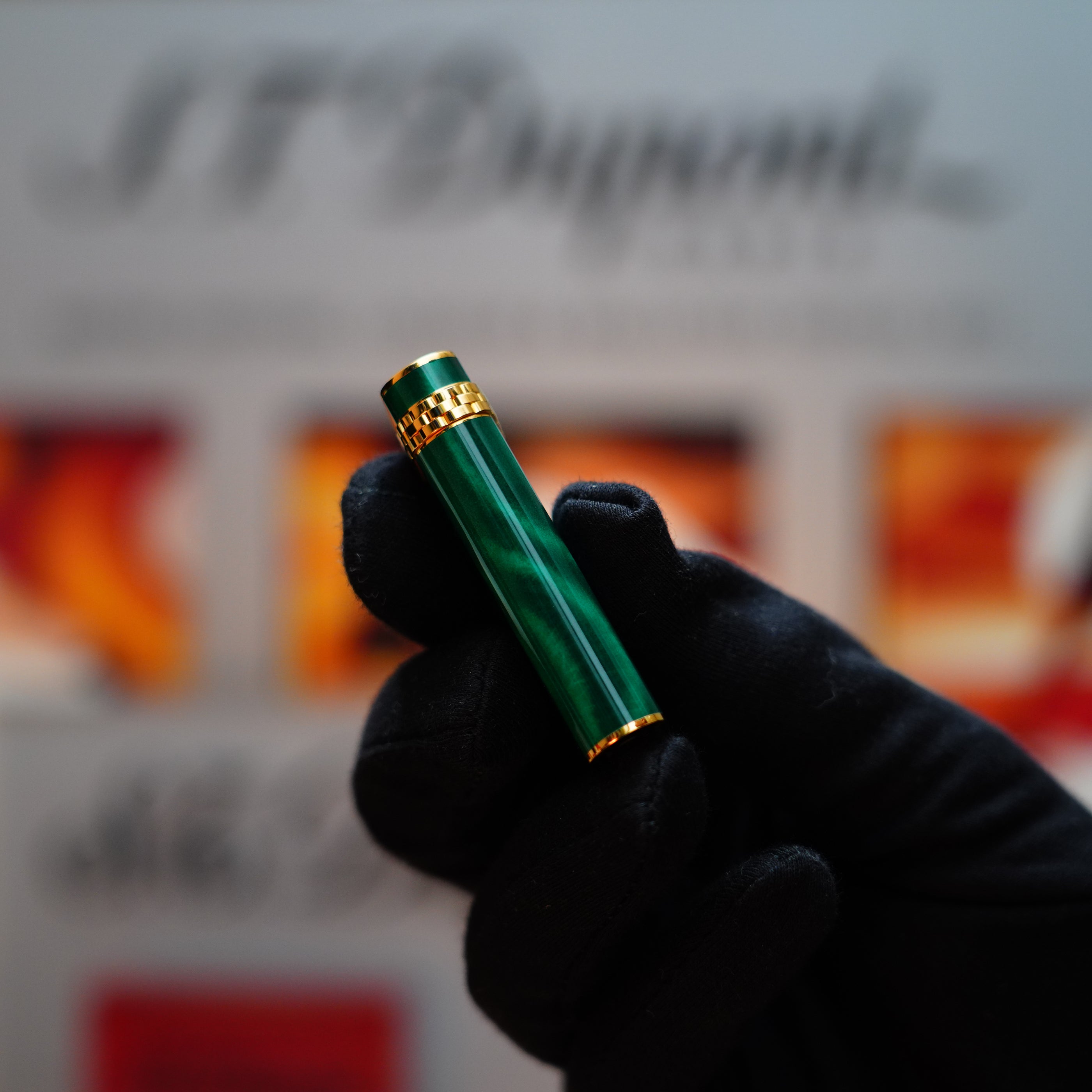 A hand carefully holds the Vintage 1989 Cartier 24k Gold Finish Rare Lacquer Malachite Gemstone Mini Lighter, its green shade echoing that of malachite gemstones, adorned with an 18k gold ring on the door, set against a softly blurred background.