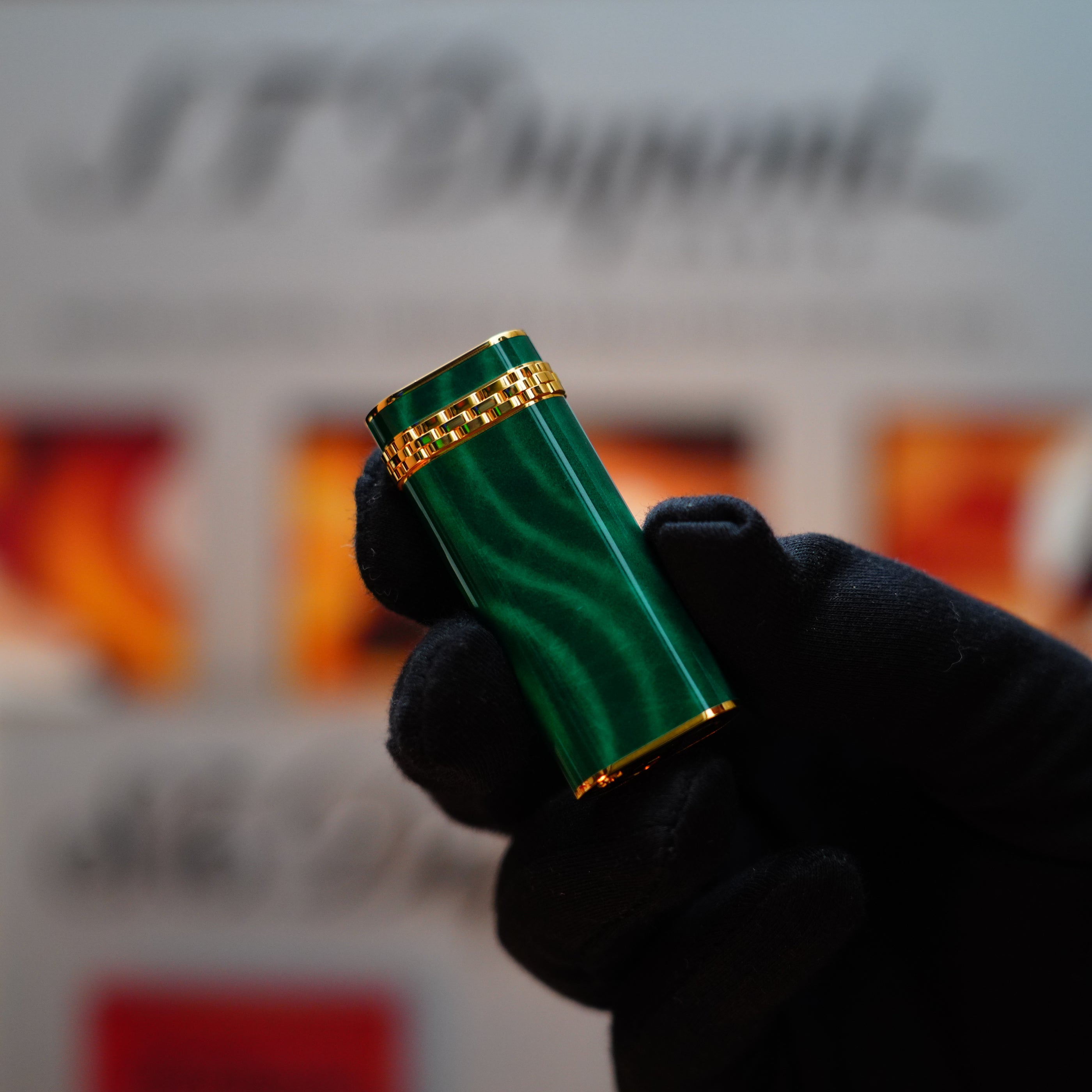 A gloved hand holds a Vintage 1989 Cartier 24k Gold Finish lighter with Rare Lacquer Malachite Gemstone and an 18k Gold Ring on the Door, framed against a blurred background featuring the S.T. Dupont logo.