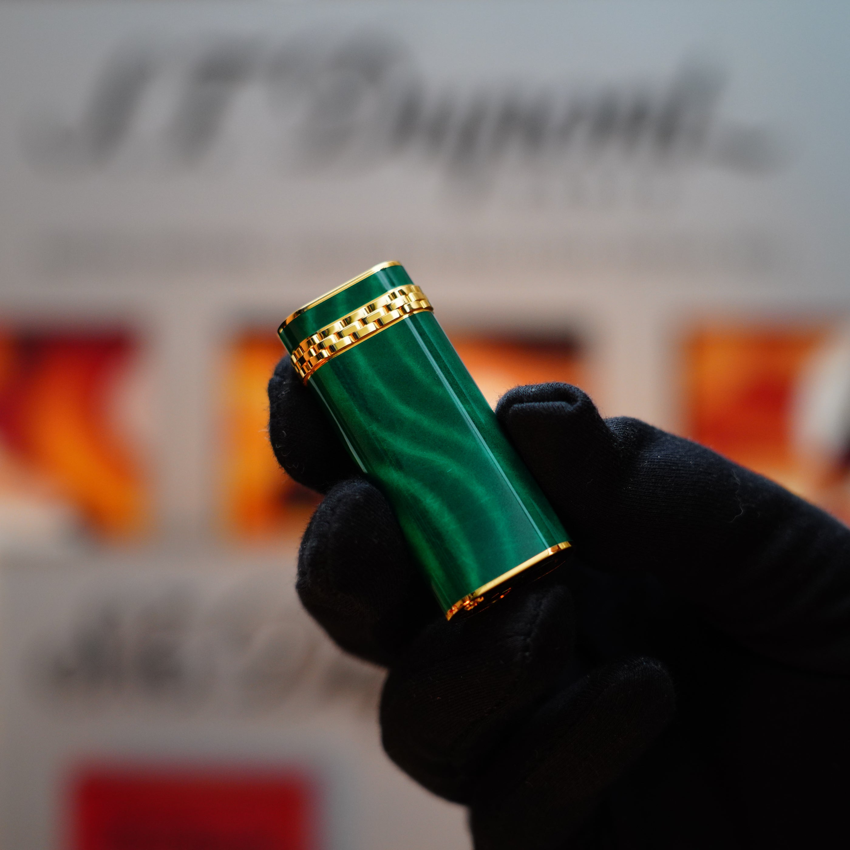 A hand in a black glove holds a Vintage 1989 Cartier lighter with green lacquer and gold trim, featuring an 18k gold ring on the door, set against a blurred background.