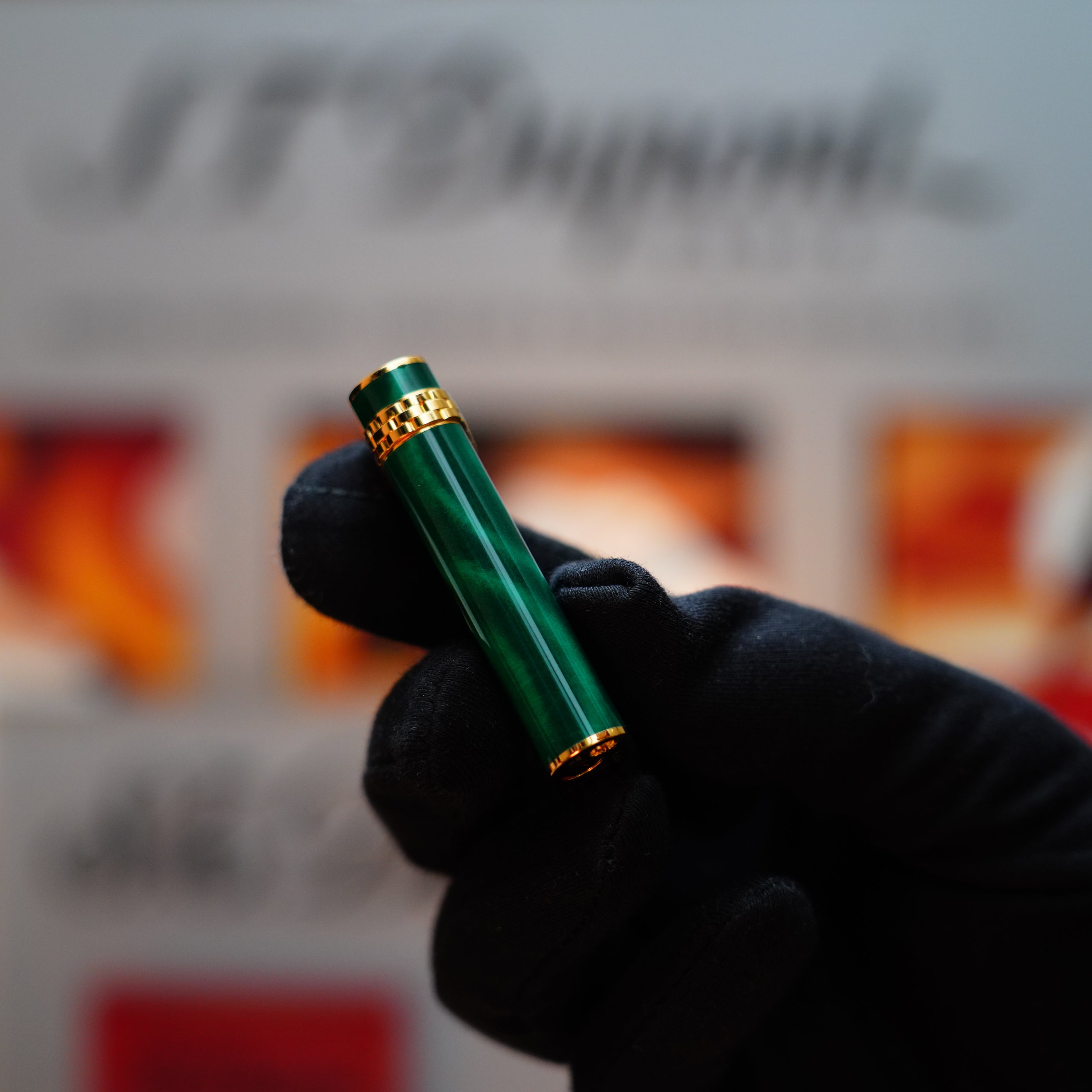 A gloved hand holds a green and gold cylindrical object, identified as the Vintage 1989 Cartier 24k Gold Finish Rare Lacquer Malachite Gemstone Mini with 18k Gold Ring on Door Lighter, against a blurred background.