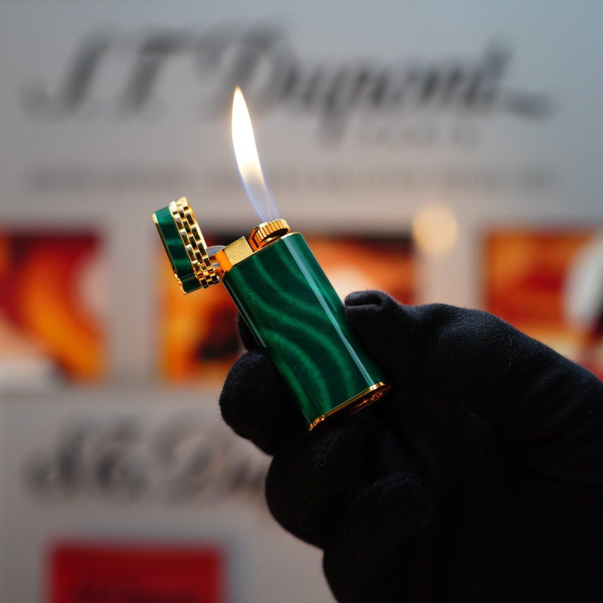 A gloved hand holds the lit Vintage 1989 Cartier Mini Lighter, featuring a 24k gold finish and rare lacquer with malachite gemstones, complete with an 18k gold ring on the door, against a blurred background.