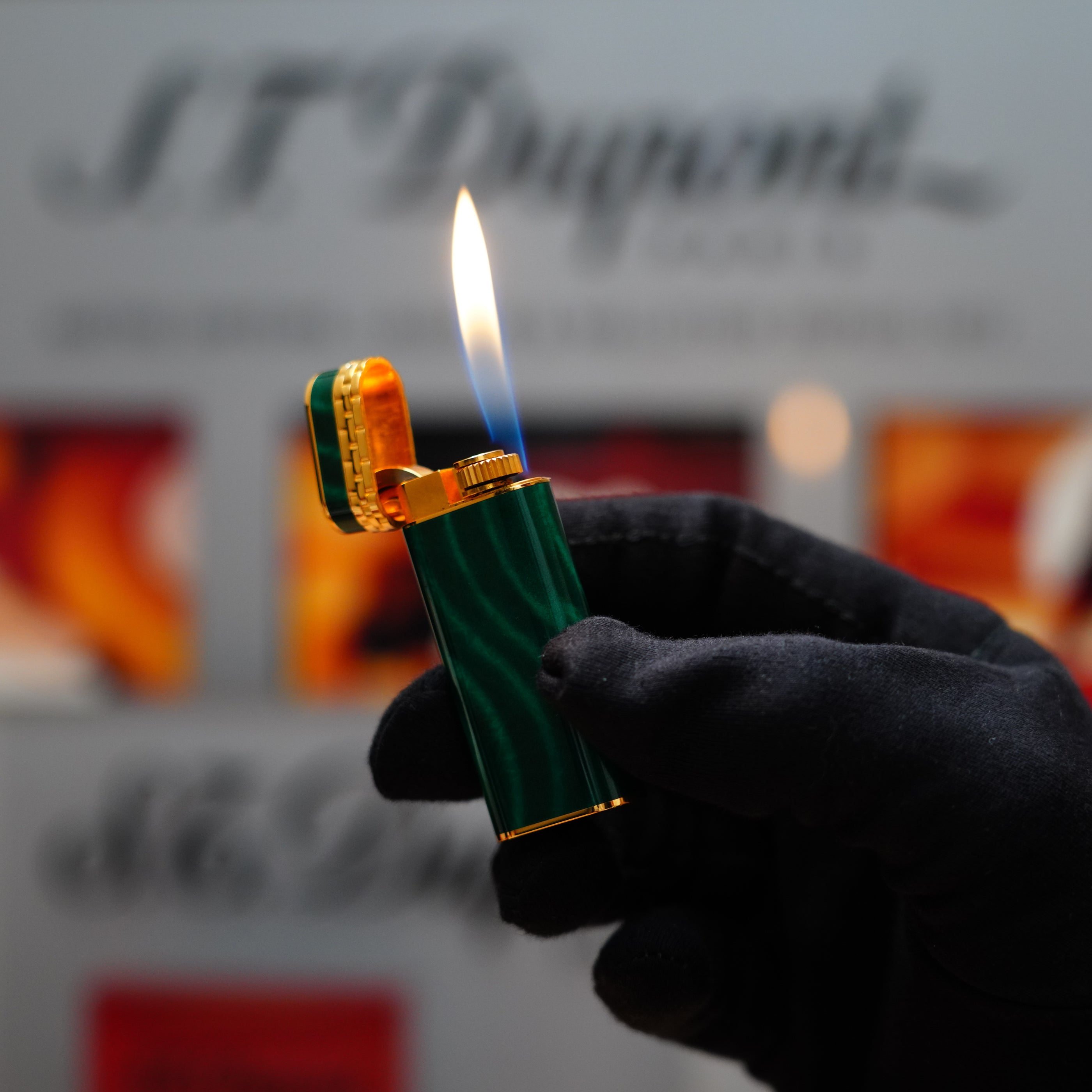 A gloved hand holds a lit Vintage 1989 Cartier 24k Gold Finish Rare Lacquer Malachite Gemstone Mini lighter, its flame dancing against a blurred background, while the 18k gold ring on its door subtly catches the light.