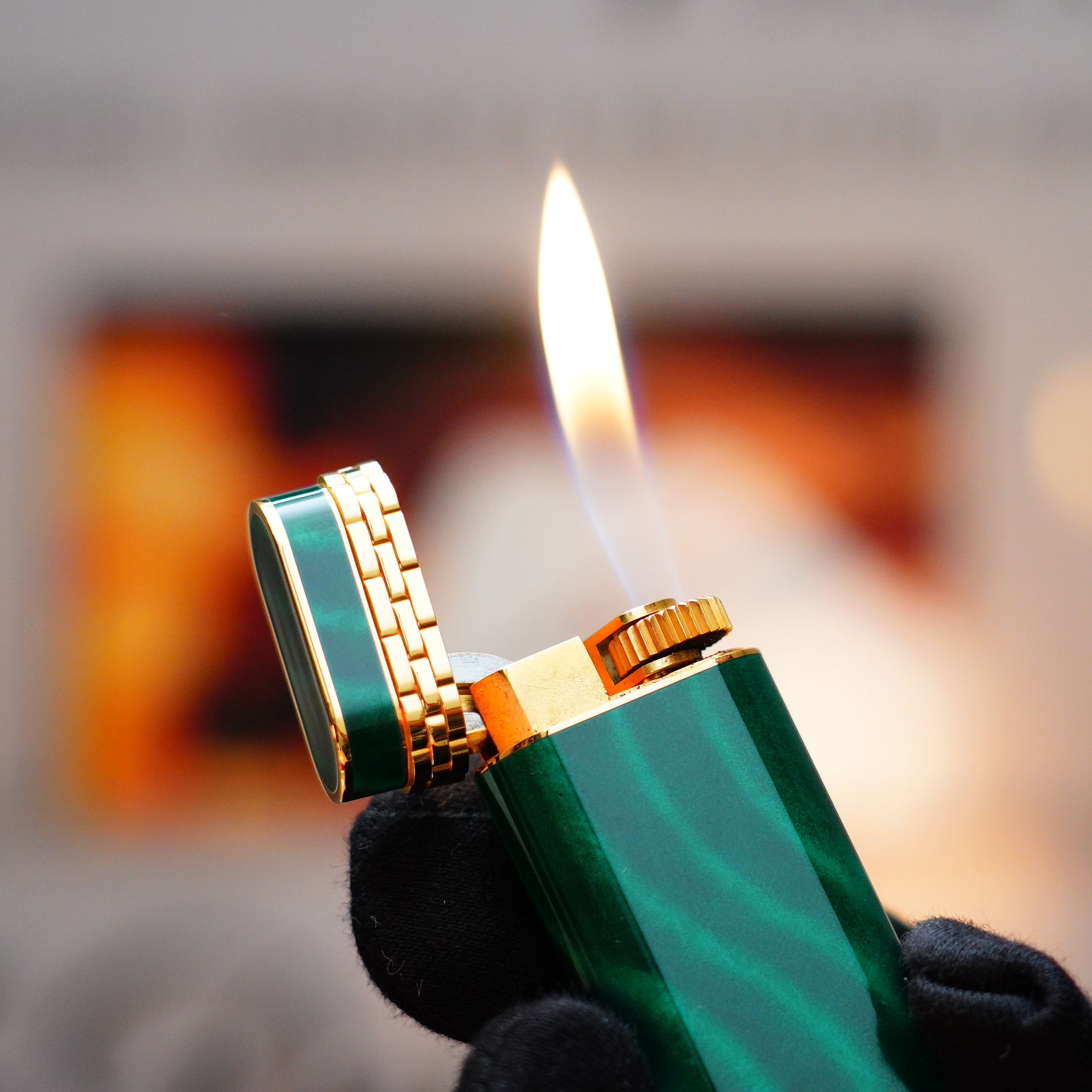 A close-up of a person holding a lit Vintage 1989 Cartier 24k Gold Finish Rare Lacquer Malachite Gemstone Mini Lighter with an 18k Gold Ring on the Door, its flame dancing against a blurred background.