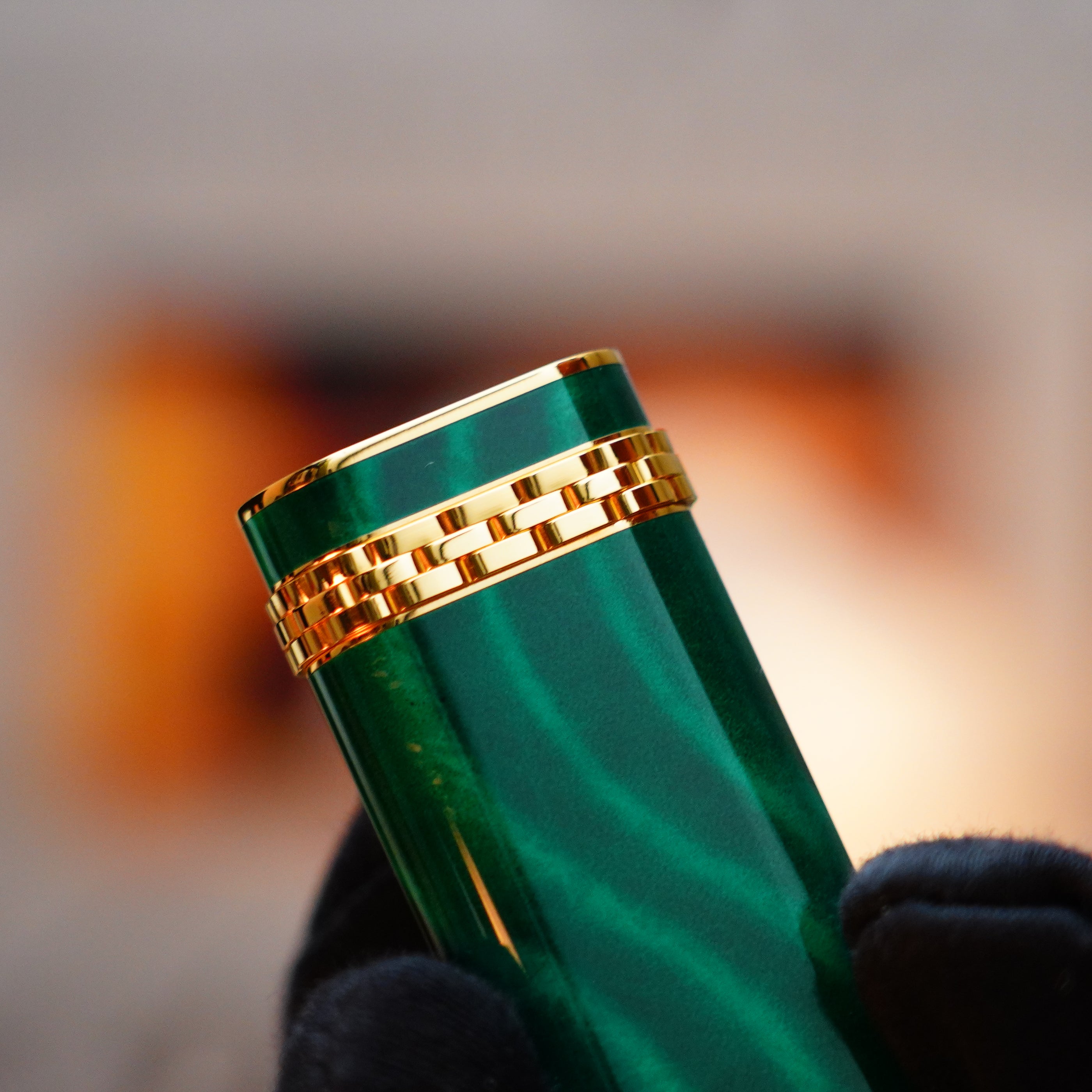 Close-up of a hand holding a cylindrical object with a green finish and an 18k gold ring, resembling the Vintage 1989 Cartier 24k Gold Finish Rare Lacquer Malachite Gemstone Mini lighter, against a blurred background.