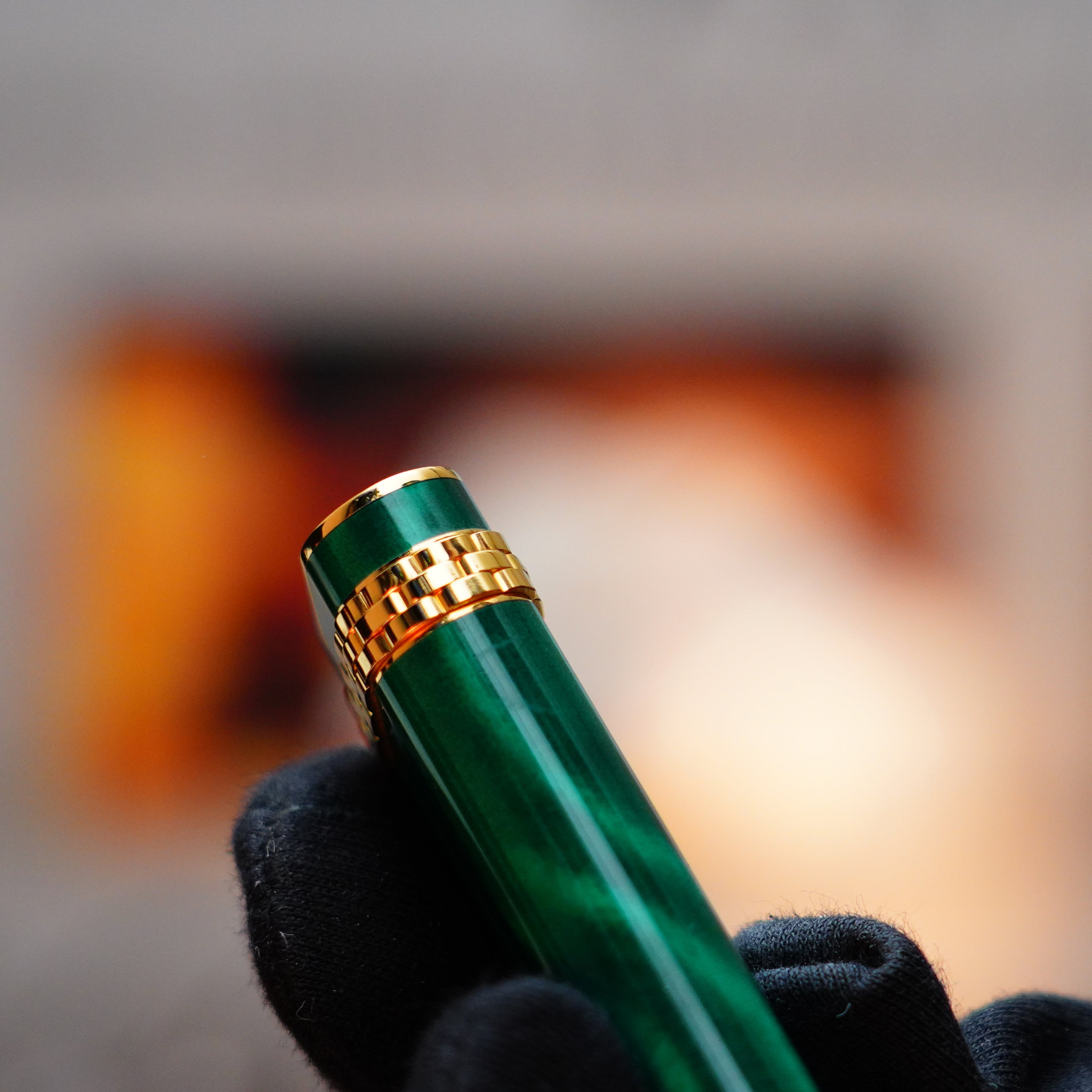 A gloved hand elegantly holds the Vintage 1989 Cartier Mini, featuring a green lacquer finish reminiscent of malachite gemstone, complemented by a 24k gold clip and an 18k gold ring, set against a softly blurred background.