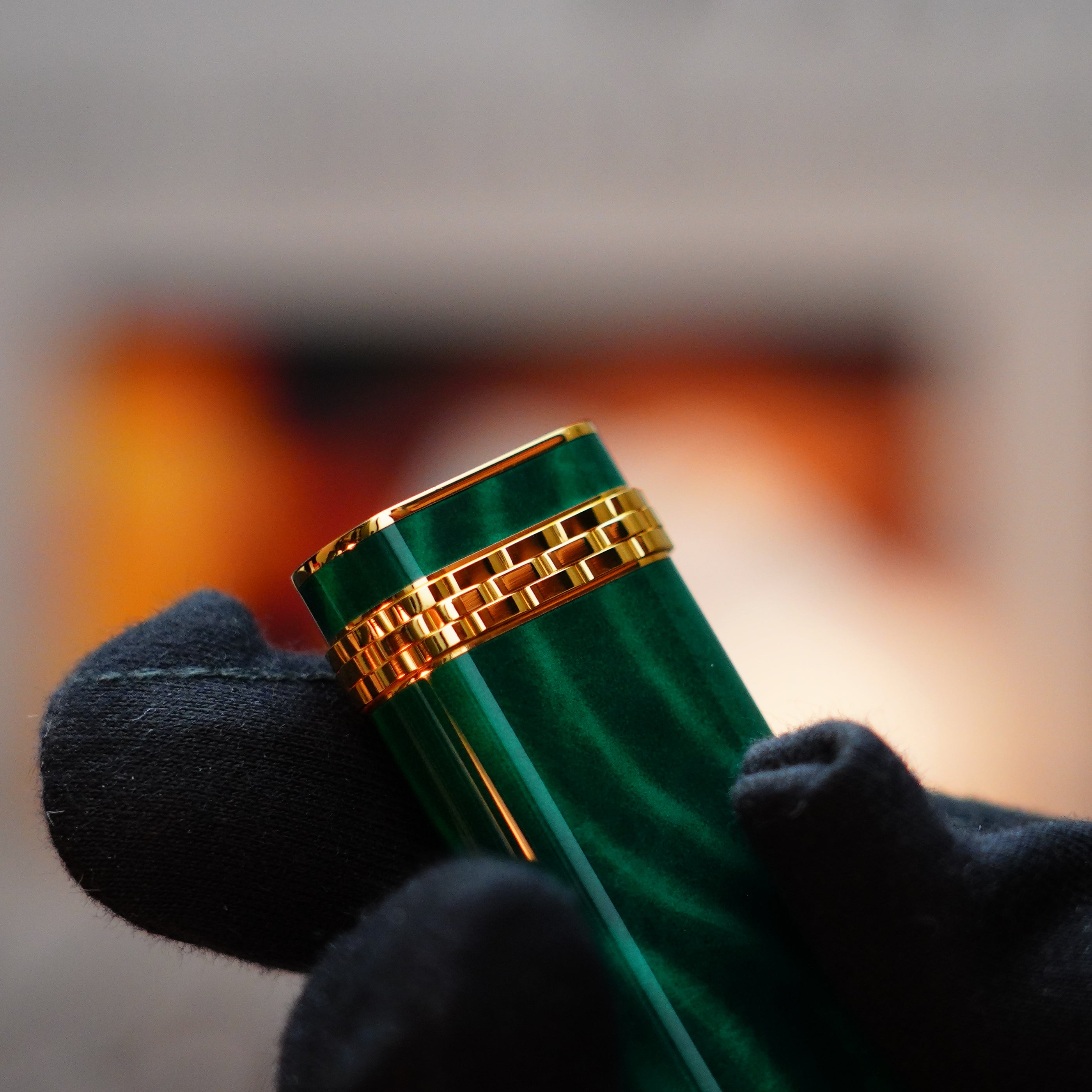 A gloved hand holds a Vintage 1989 Cartier 24k Gold Finish Rare Lacquer Malachite Gemstone Mini Lighter, its green form adorned with an 18k gold ring on the door, against a blurred background.