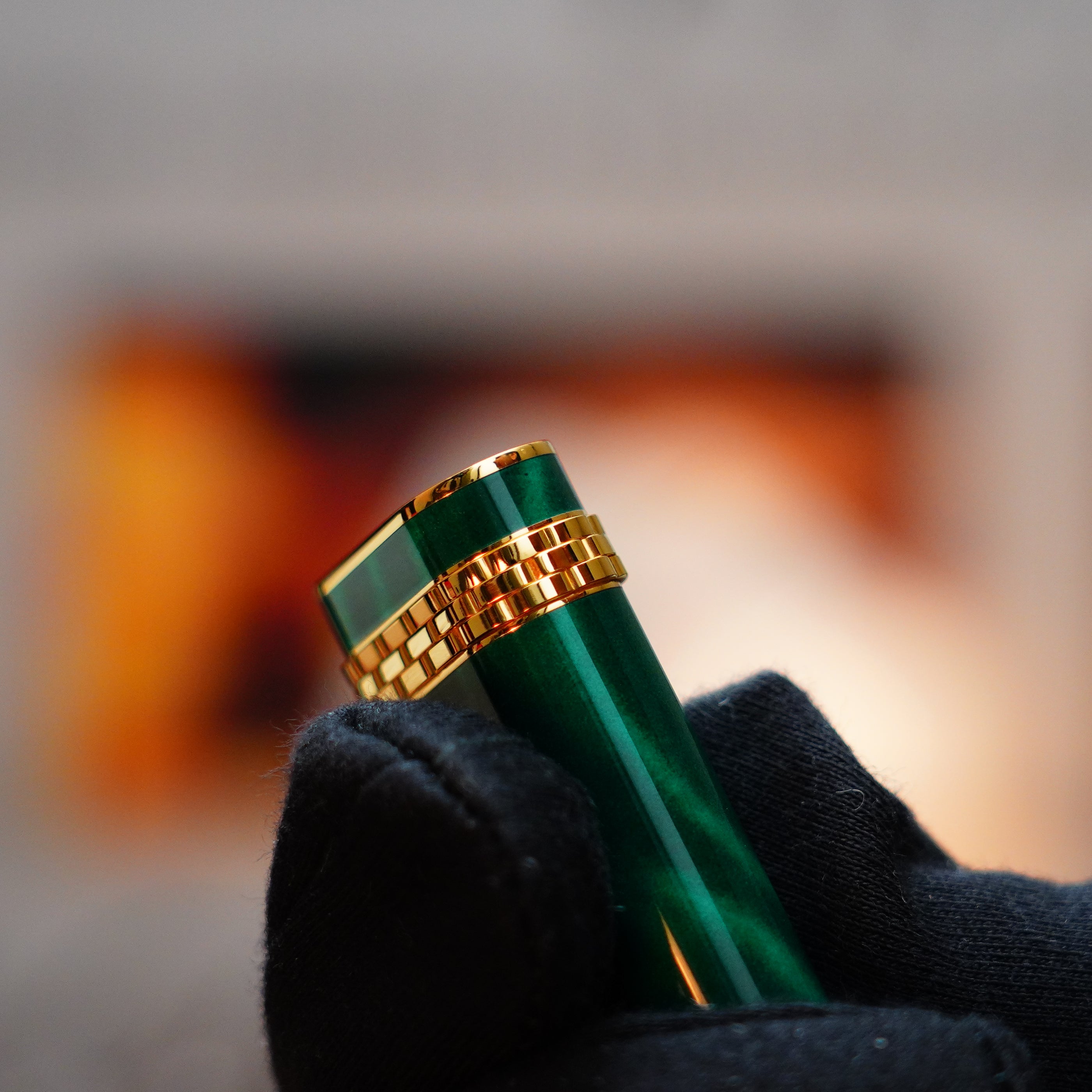 A gloved hand holds a vintage 1989 Cartier lighter, the 24k gold-finished mini adorned with rare lacquer malachite gemstone and an 18k gold ring on the door, as a flickering flame dances in the background.