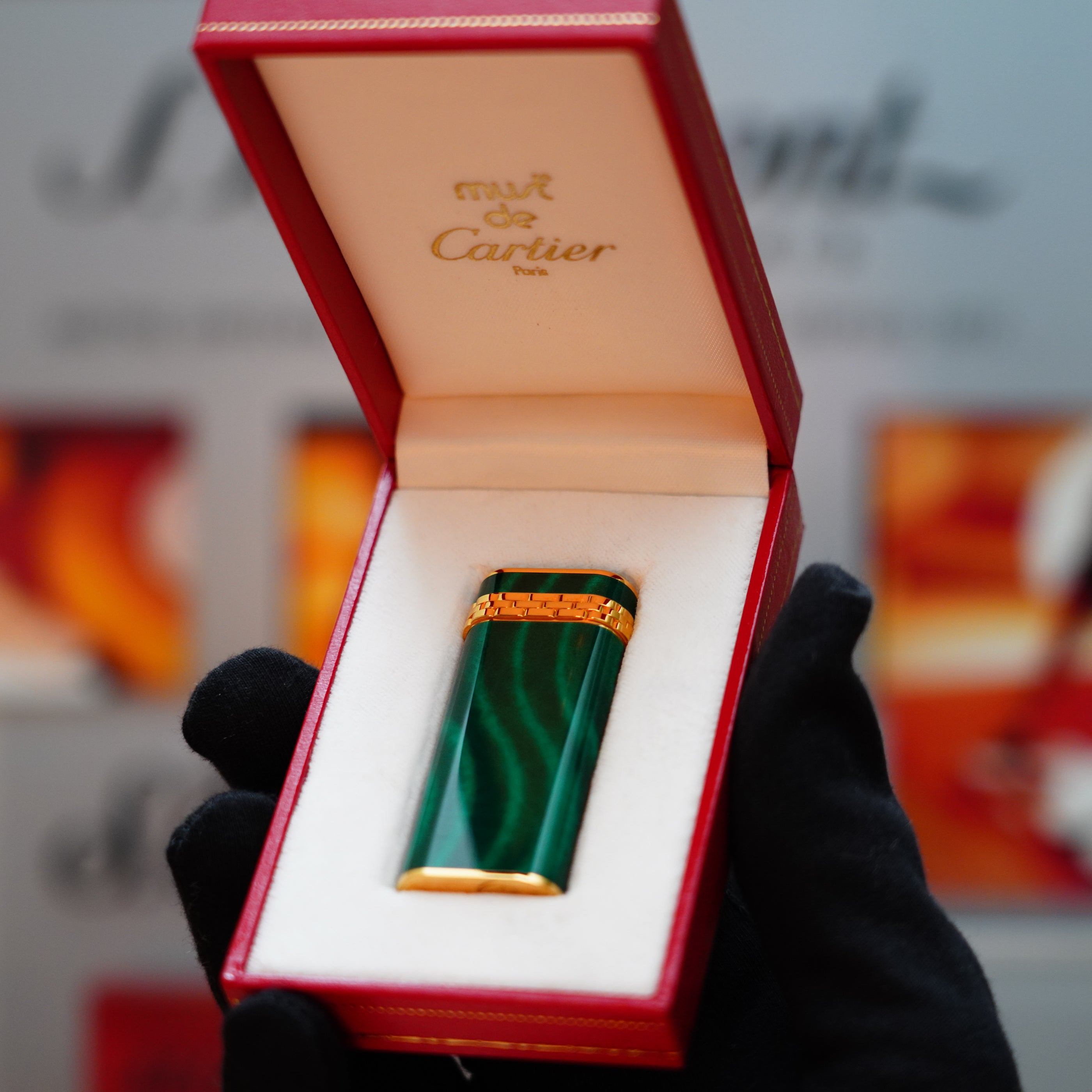 A gloved hand holds an open red box, revealing a Vintage 1989 Cartier lighter with a 24k gold finish and a malachite gemstone, featuring the iconic Cartier branding inside.