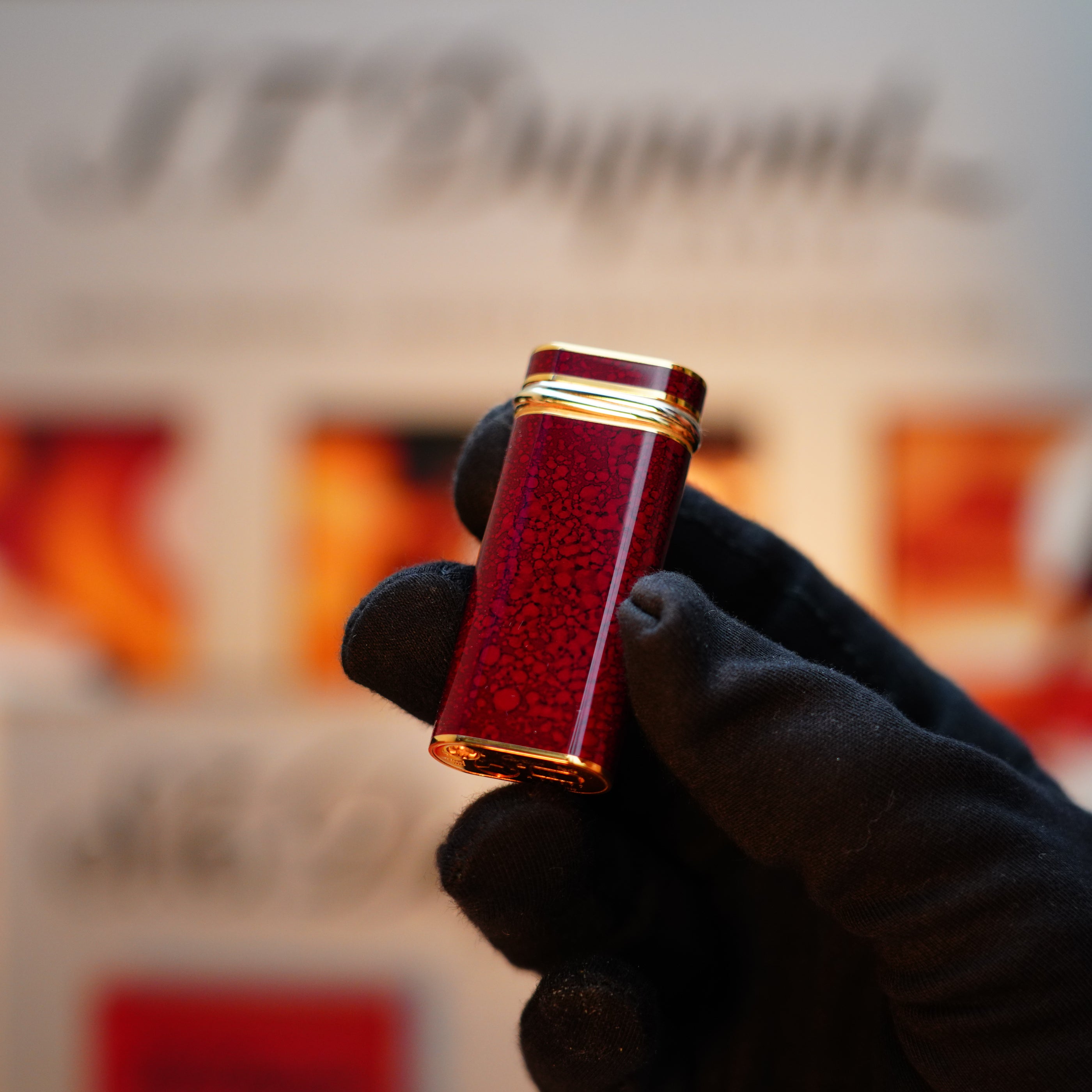 A gloved hand holds a Vintage 1989 Cartier lighter, the red lacquer and 24k gold finish shimmering against a blurred backdrop of indistinct text and images, its door adorned with an 18k Trinity Ring.