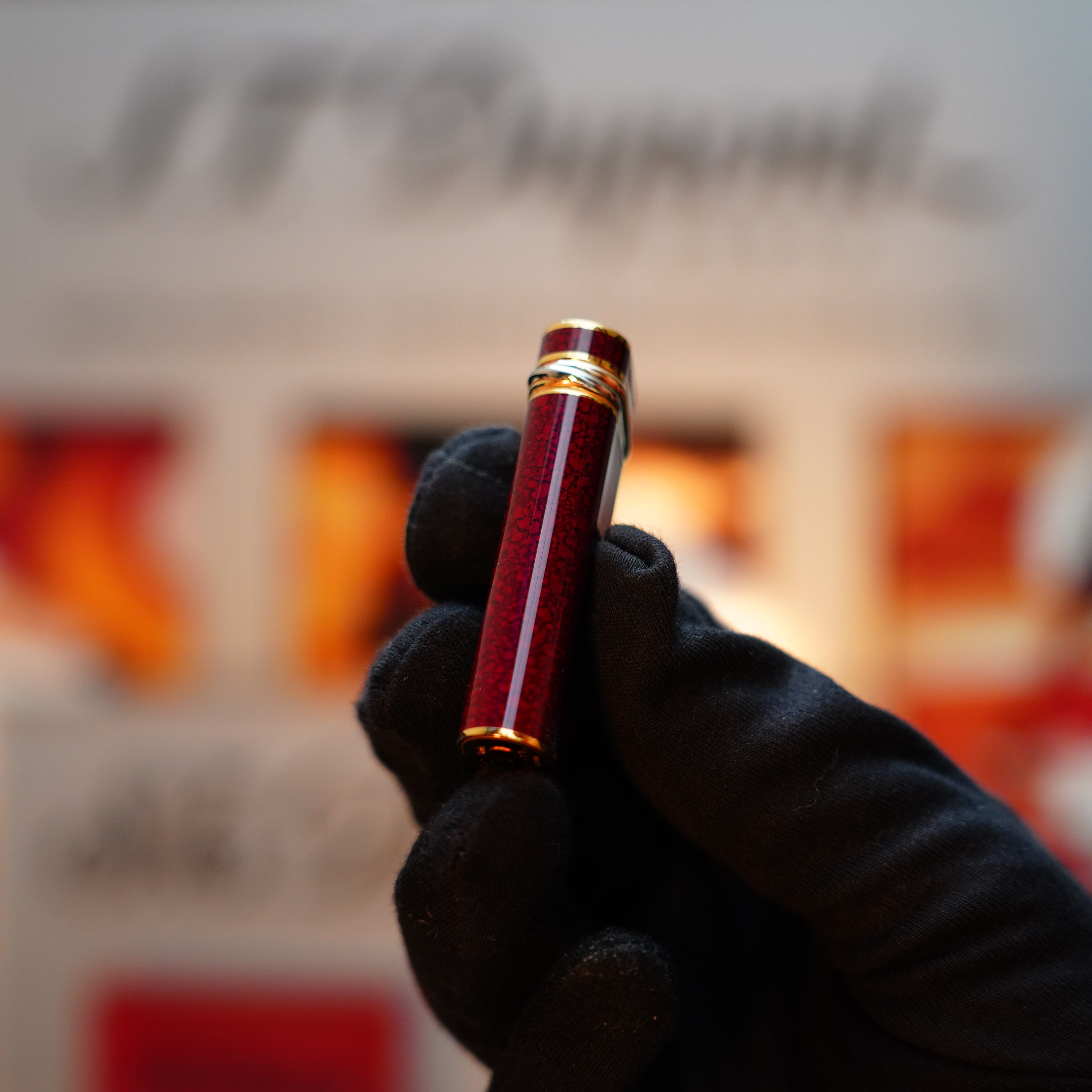 A gloved hand elegantly holds the Vintage 1989 Cartier red fountain pen with a 24k gold finish, beautifully highlighted against a blurred backdrop of branded materials and an exquisite Jasper Gemstone Mini.