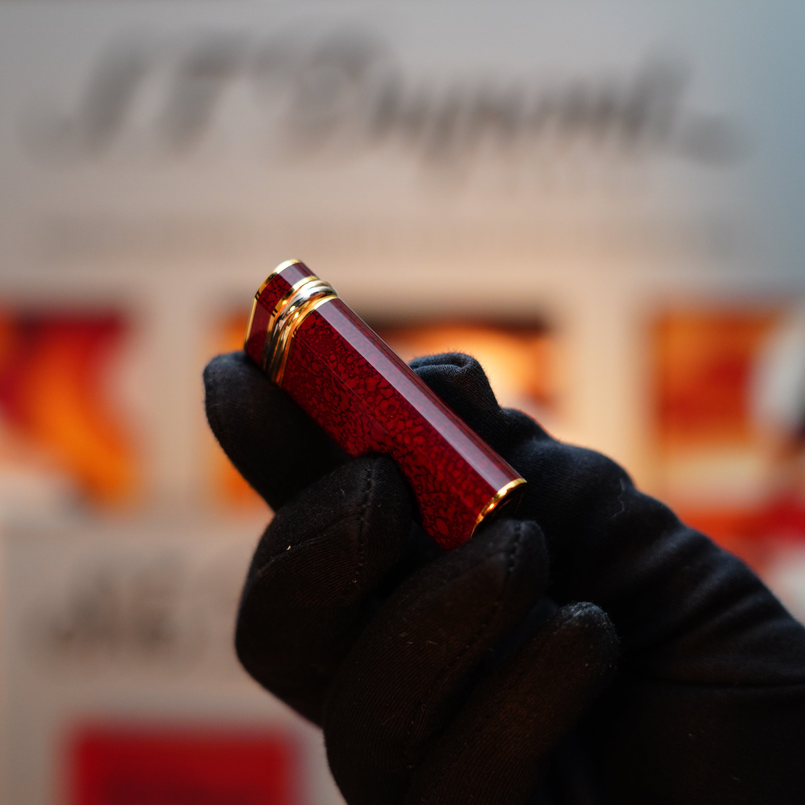 A gloved hand elegantly holds a Vintage 1989 Cartier lighter, its 24k gold finish and rare lacquer gleaming against a blurred background, accented by an 18k Trinity Ring on the door.