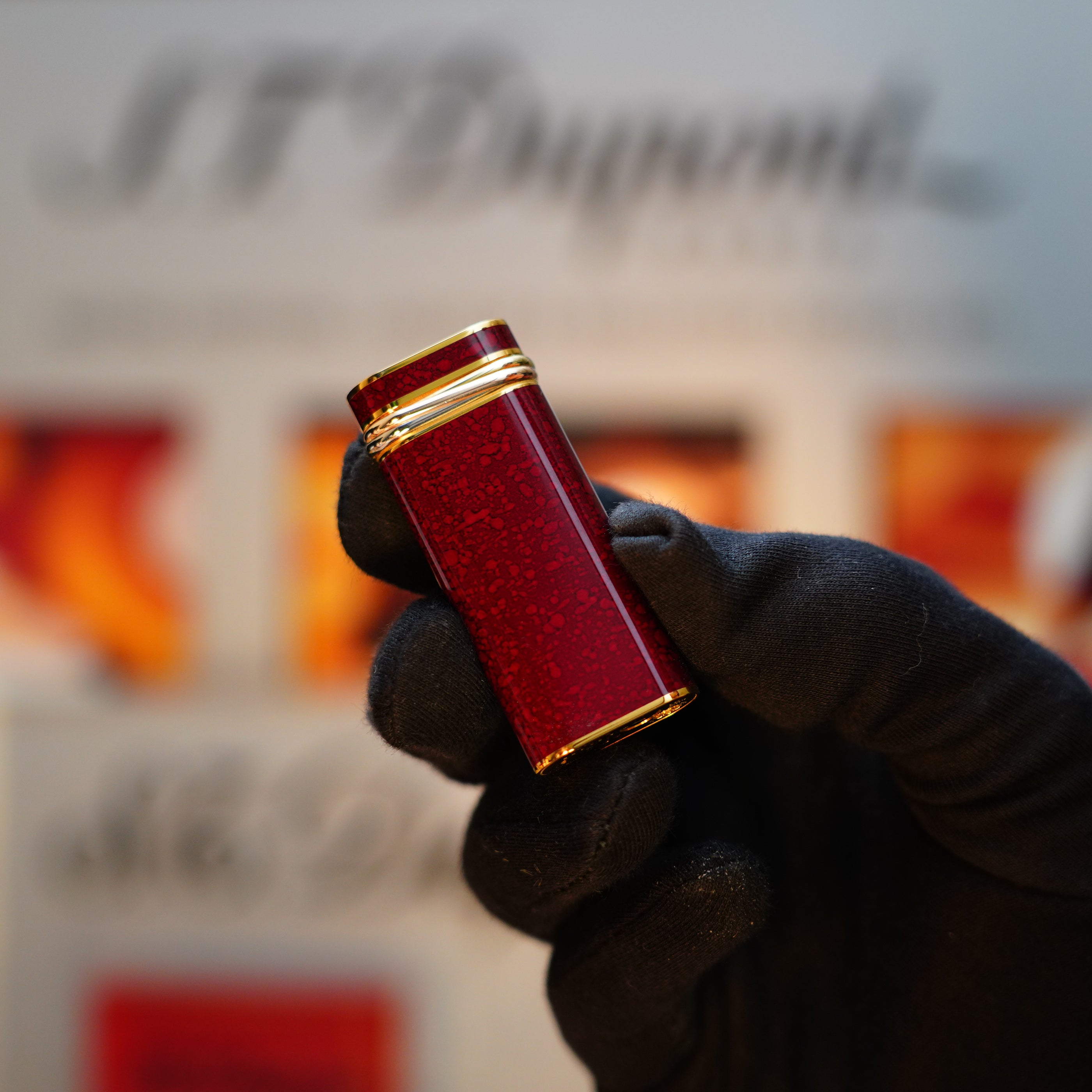 A gloved hand holds a Vintage 1989 Cartier Lighter, featuring a rare lacquer finish in red and 24k gold highlights, adorned with an 18k Trinity Ring on the door, in front of a blurred background.