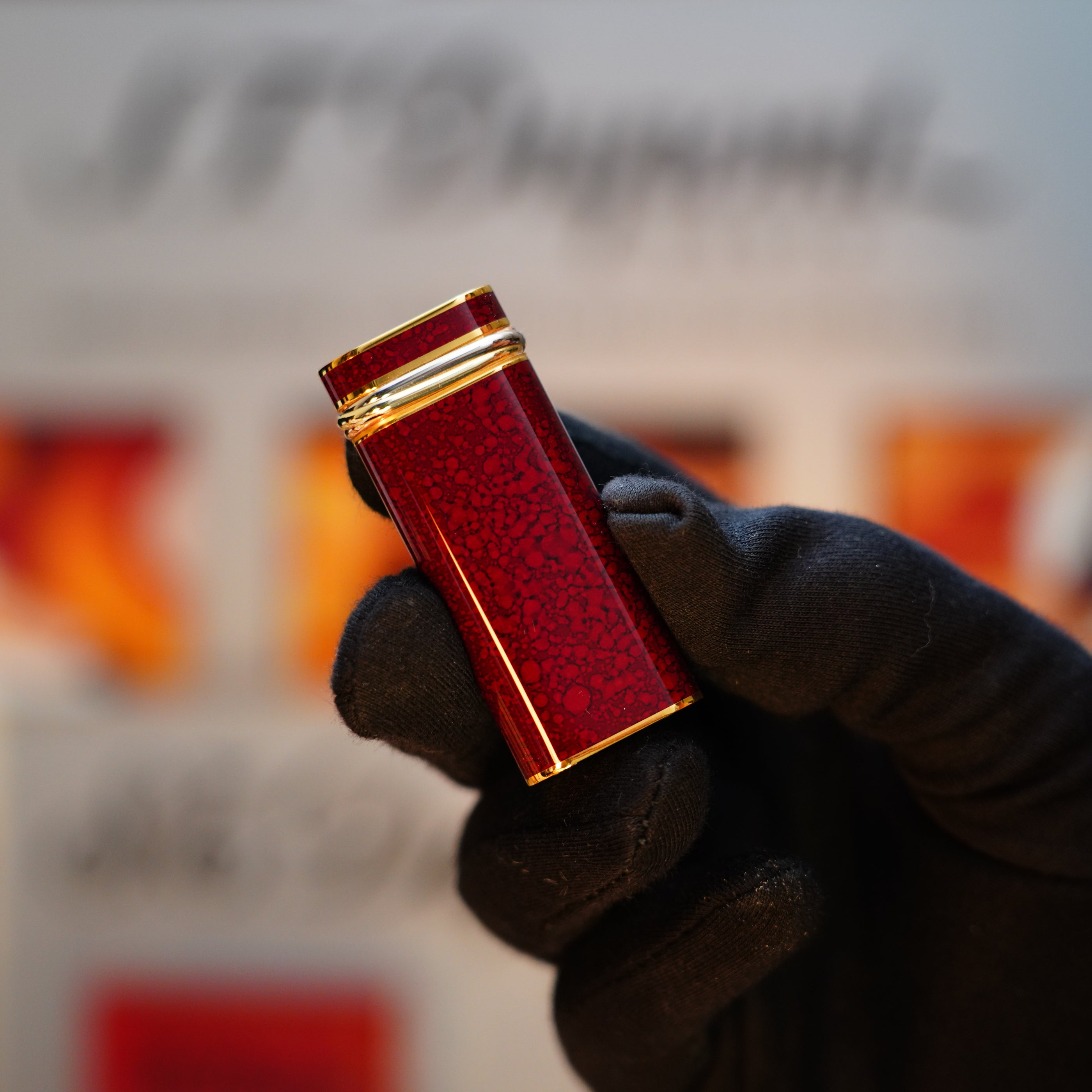 A person wearing a black glove holds a compact, red rectangular object with gold accents, resembling the Vintage 1989 Cartier 24k Gold Finish Rare Lacquer Jasper Gemstone Mini with an 18k Trinity Ring on the door lighter.