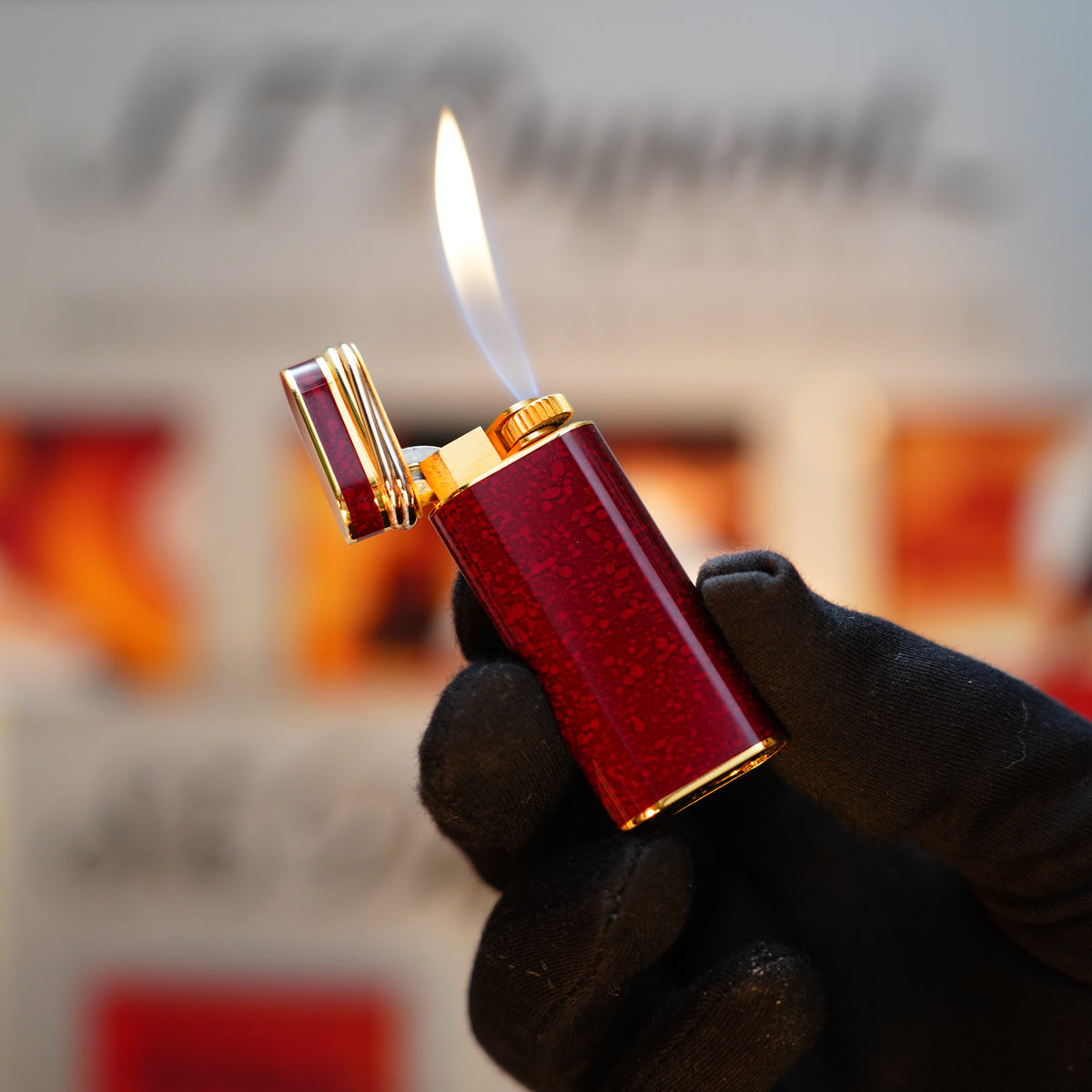 A person wearing a black glove holds a lit flame with the red and gold Vintage 1989 Cartier 24k Gold Finish Rare Lacquer Jasper Gemstone Mini with an 18k Trinity Ring on Door Lighter. The blurred background displays faint text and colors.