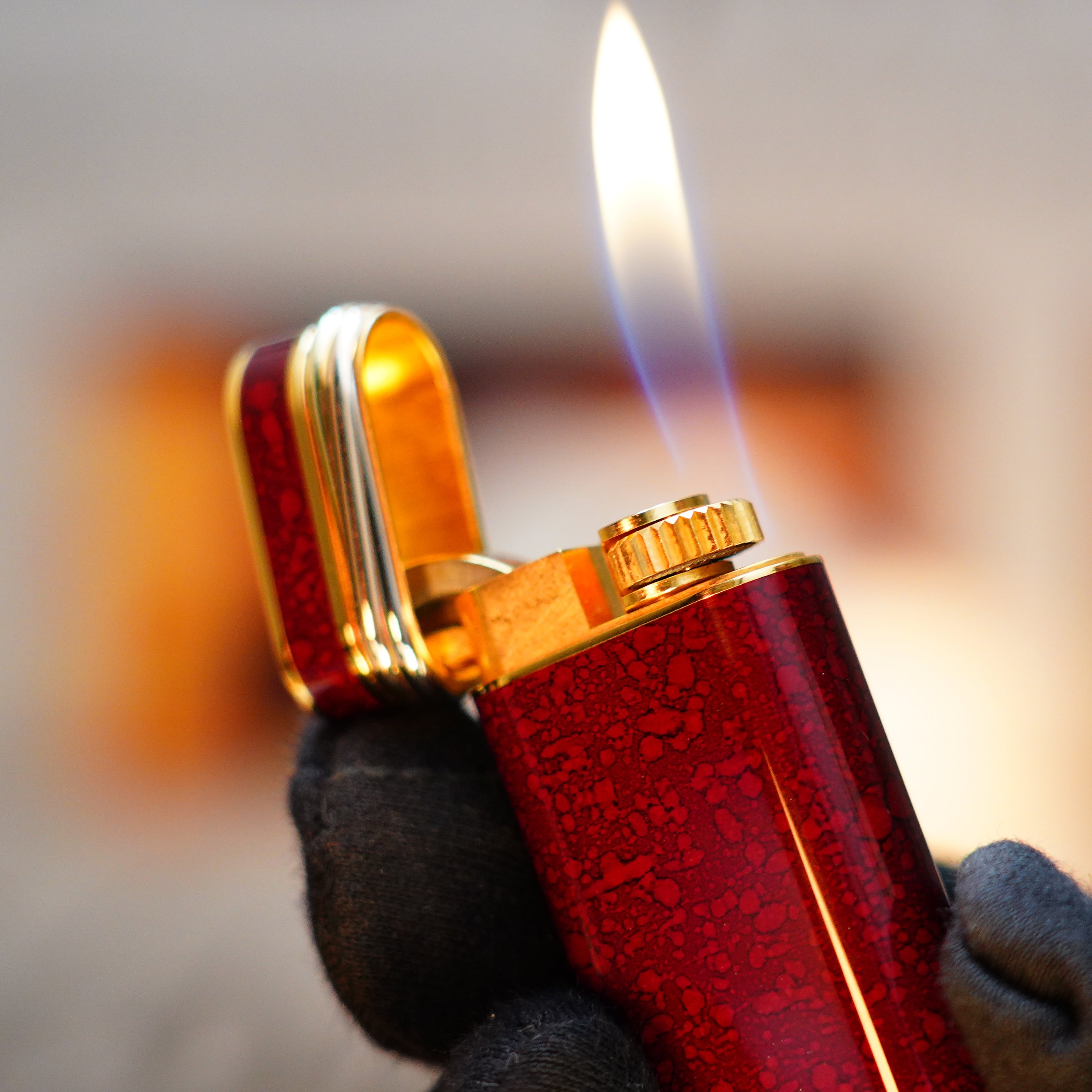 A gloved hand holds an open Vintage 1989 Cartier lighter with a 24k gold finish, adorned with a rare lacquer and jasper gemstone design, complete with an 18k Trinity ring on the door, producing a flame.