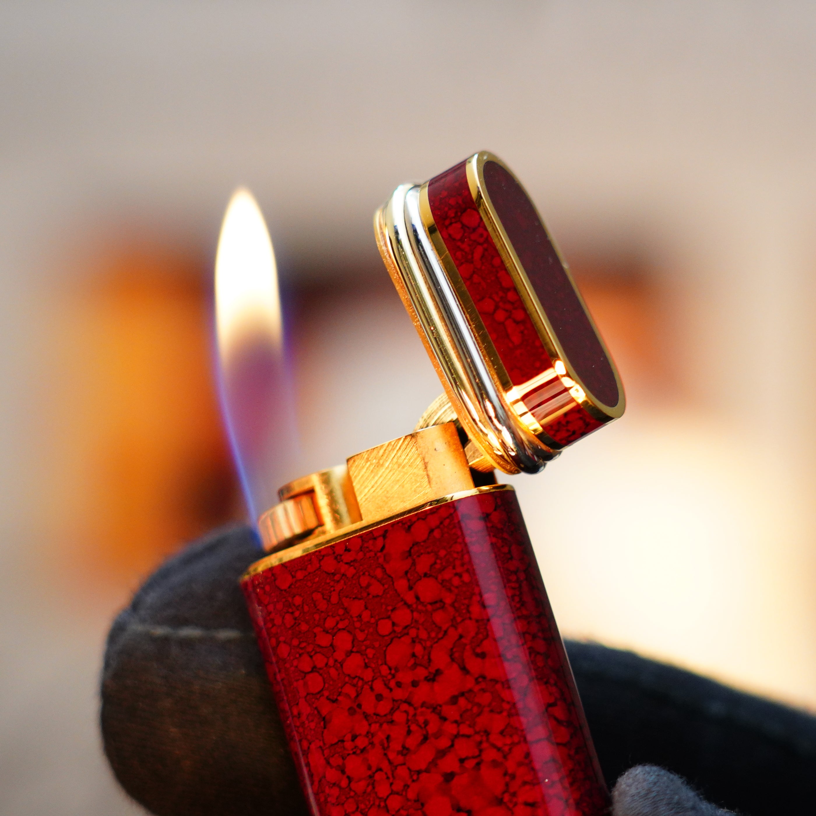 A gloved hand holding a lit Vintage 1989 Cartier lighter with a blue flame, featuring a 24k gold finish and rare lacquer Jasper gemstone, complete with an 18k Trinity ring on the door.