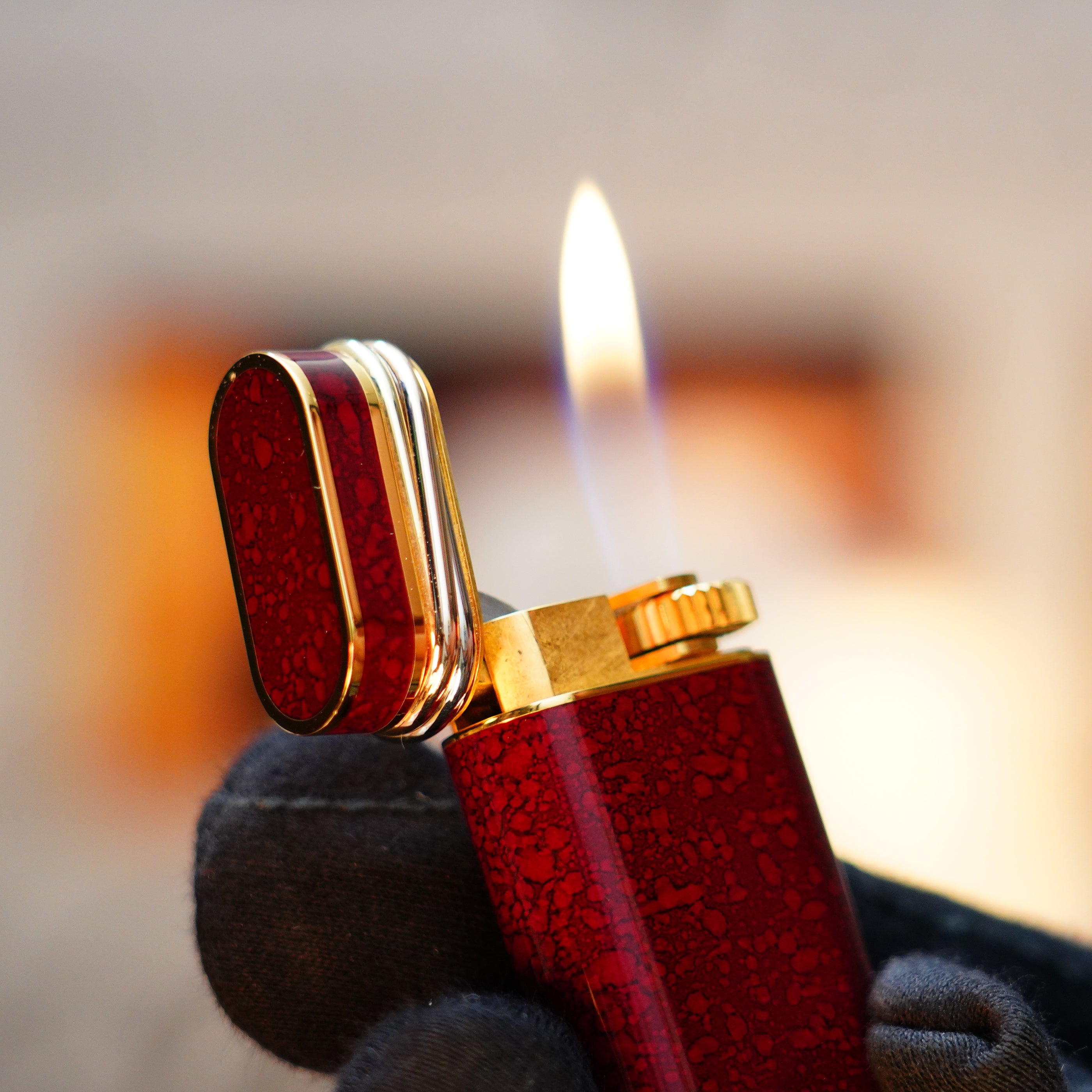 A black-gloved hand elegantly holds a Vintage 1989 Cartier 24k Gold Finish Rare Lacquer Jasper Gemstone Mini Lighter, its red and gold casing gleaming as the flame flickers warmly, complete with an 18k Trinity Ring on the door.