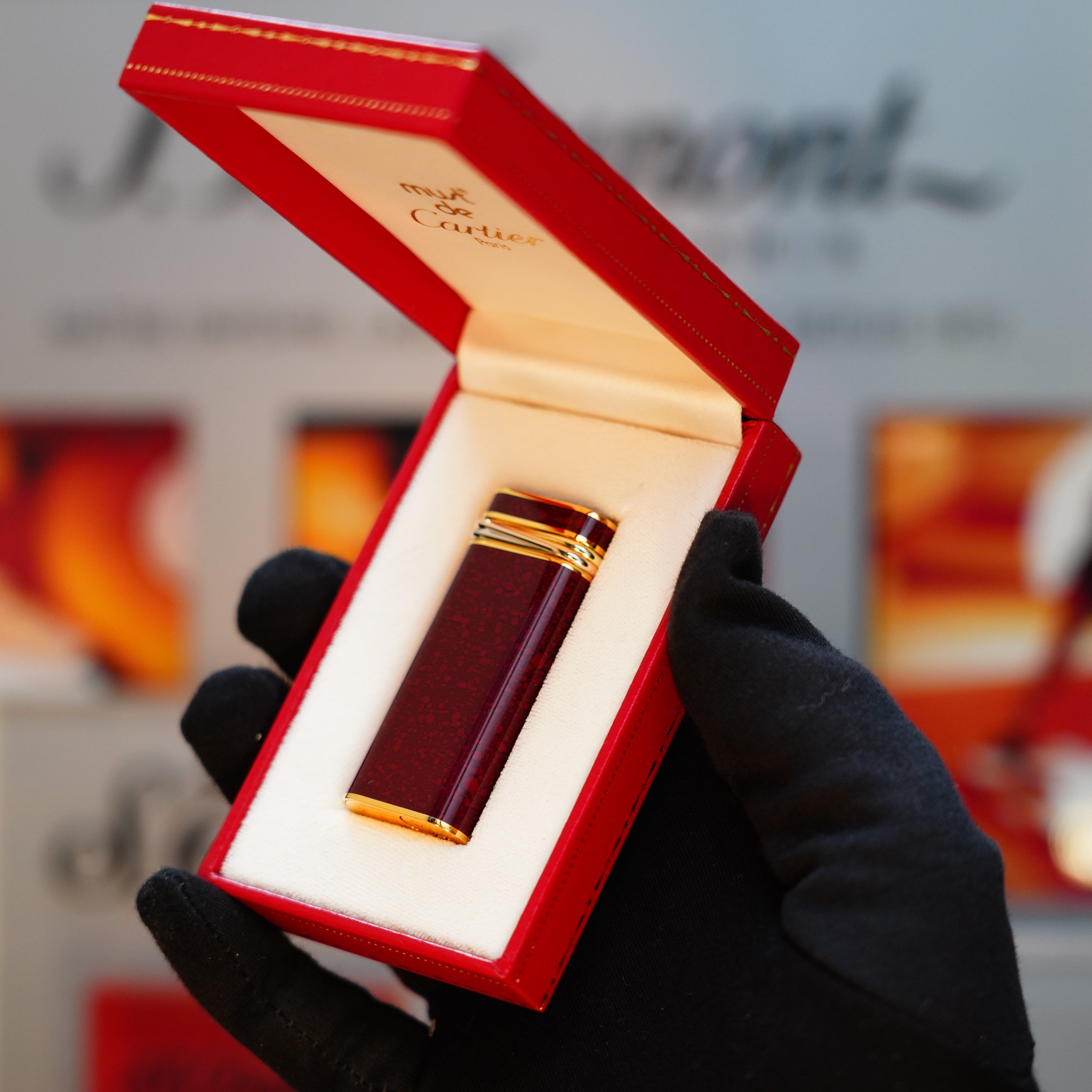 A gloved hand delicately presents an open red Cartier box, unveiling a Vintage 1989 Cartier lighter adorned with a 24k gold finish and rare lacquer, featuring a Jasper gemstone mini and an 18k Trinity ring on the door, exuding timeless elegance.