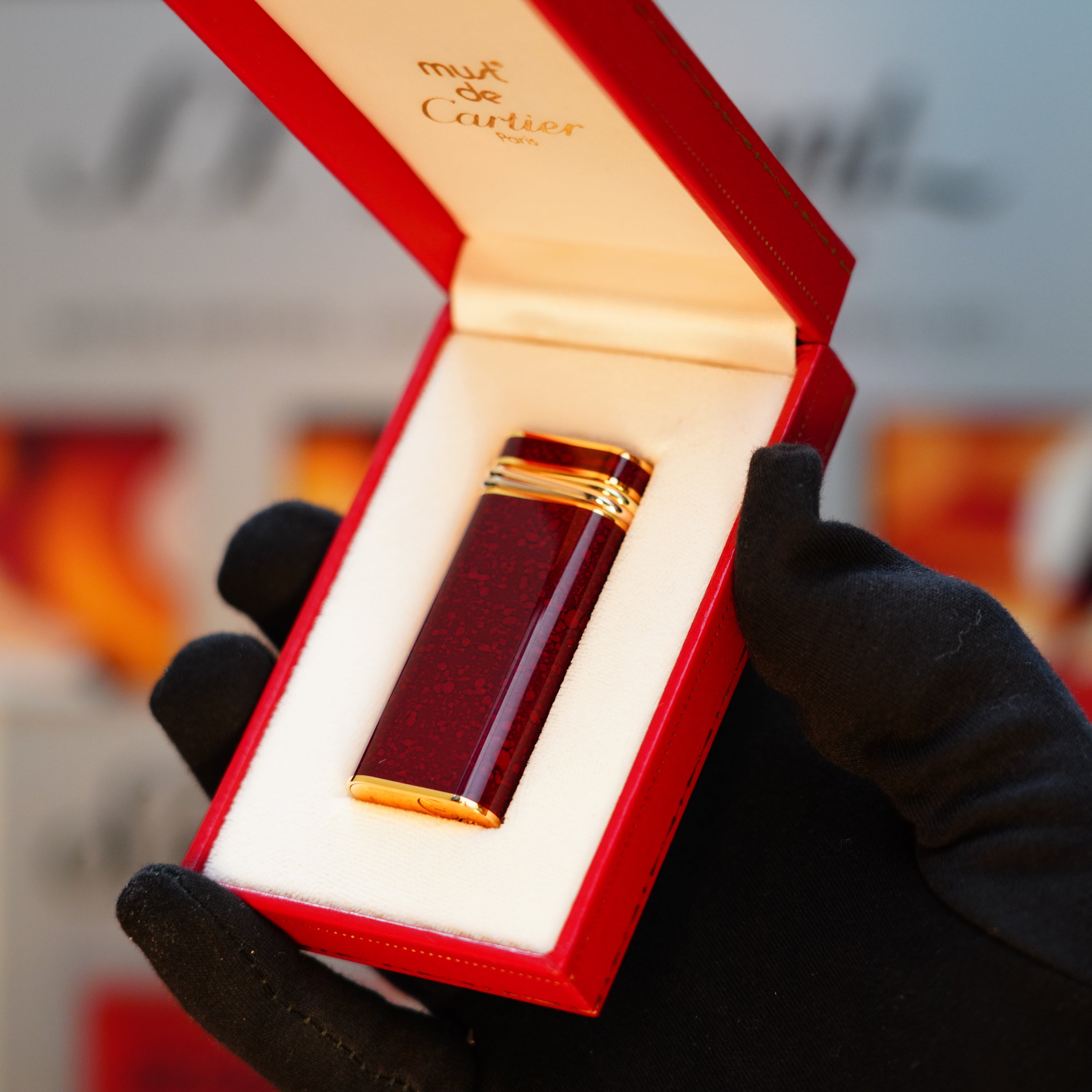 A red and gold perfume bottle rests in an open red box, elegantly cradled by a hand wearing a black glove, evoking the luxurious allure of a Vintage 1989 Cartier 24k Gold Finish Lacquer Jasper Gemstone Mini Lighter with an 18k Trinity Ring on the Door.