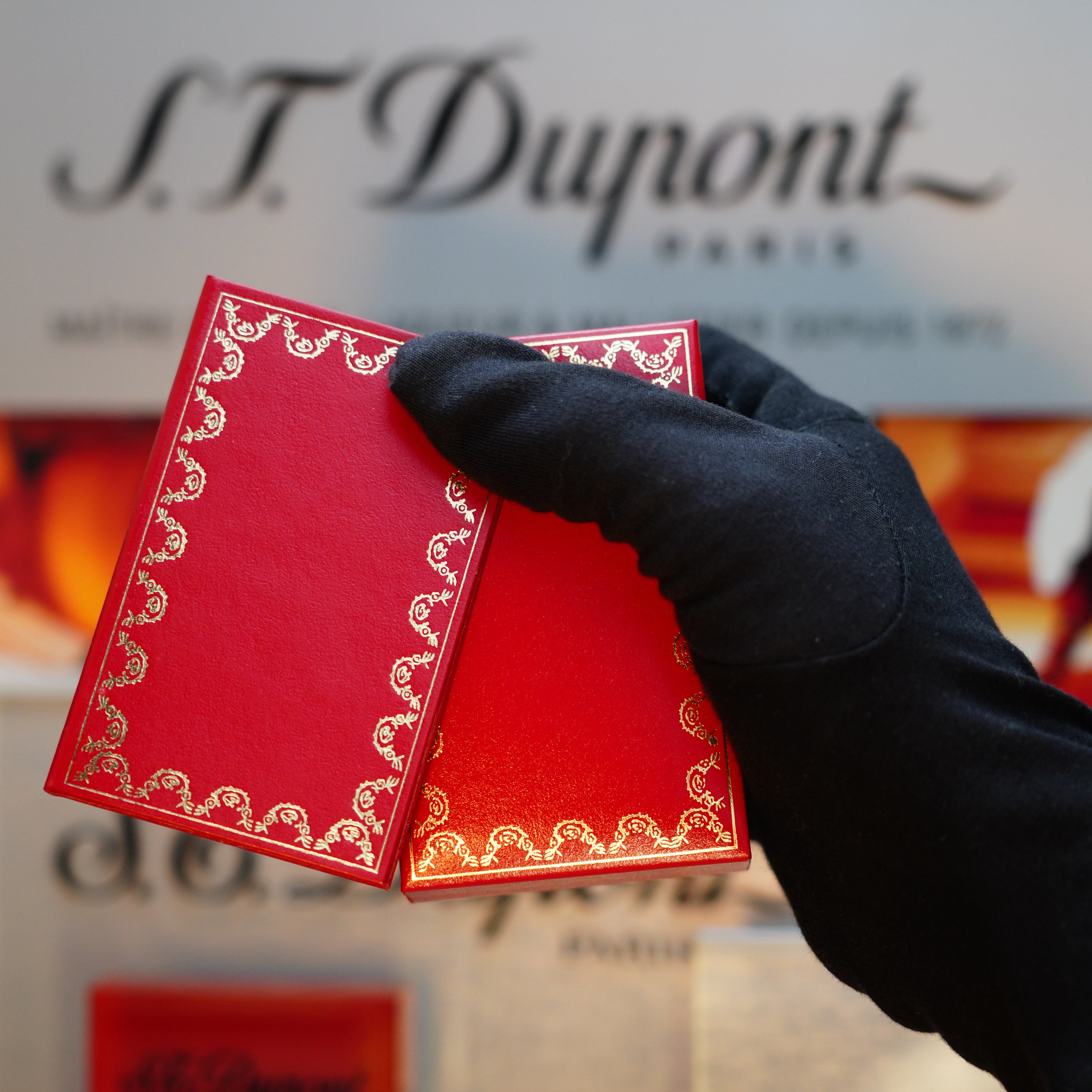 A gloved hand holds a red and gold ornate box in front of a blurred S.T. Dupont Paris sign, its elegance reminiscent of the Vintage 1989 Cartier 24k Gold Finish Rare Lacquer Jasper Gemstone Mini Lighter with an 18k Trinity Ring on the Door.