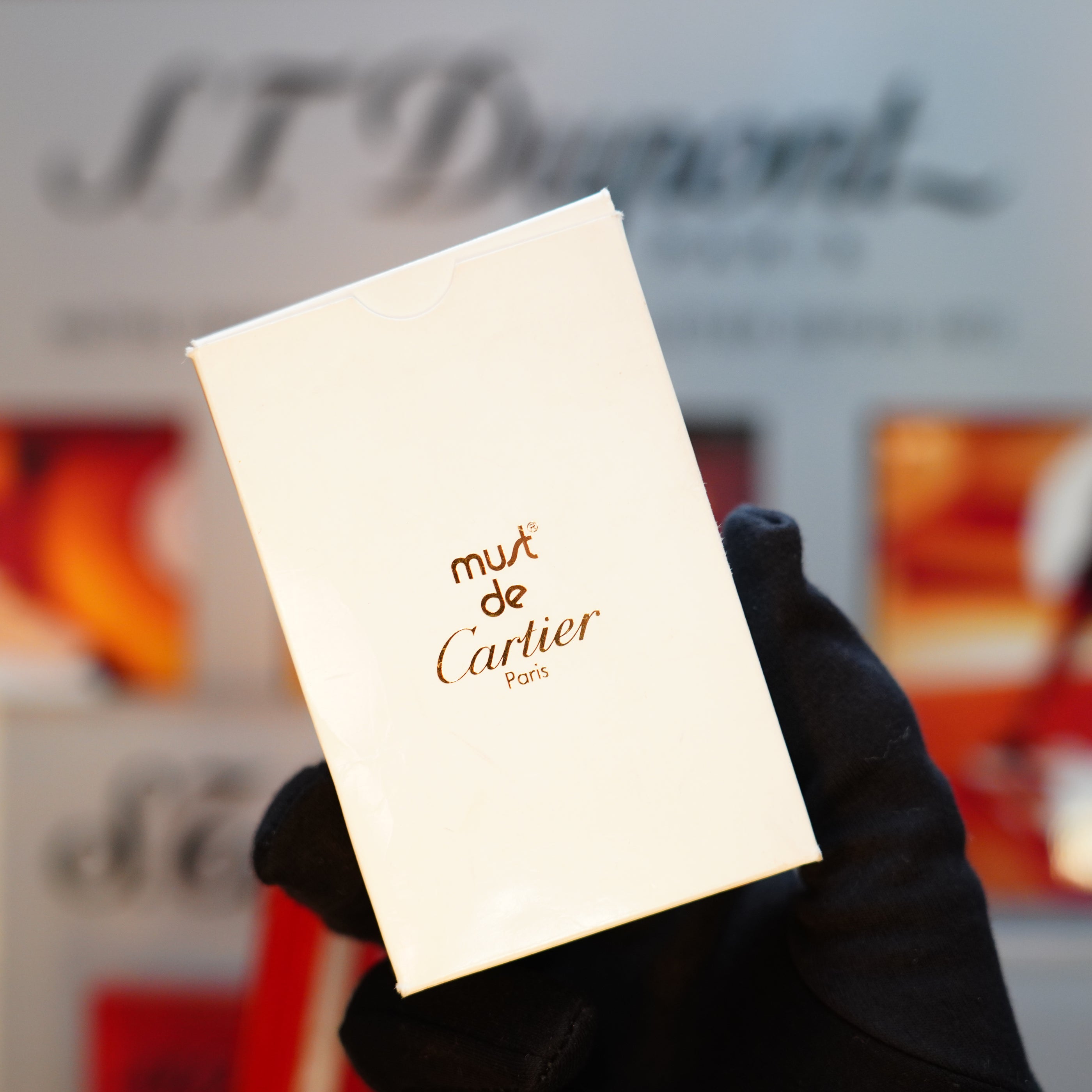 A gloved hand holds a white box labeled "Must de Cartier Paris" against a blurred background, evoking the charm of discovering a Vintage 1989 Cartier 24k Gold Finish Rare Lacquer Jasper Gemstone Mini Lighter, adorned with an elegant 18k Trinity Ring on its door.