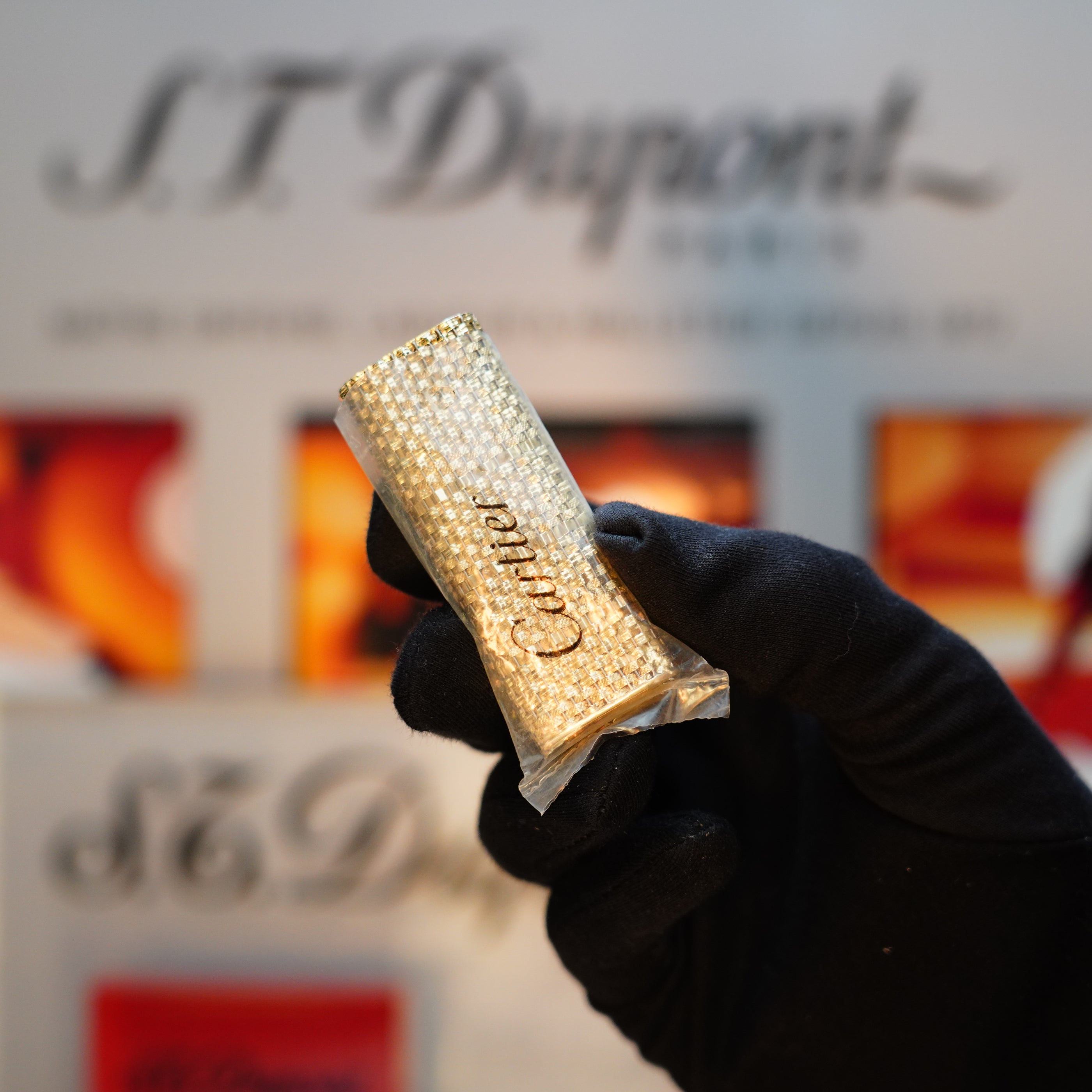 A gloved hand holds a Vintage 1980 Cartier Solid 18k Gold Jacket Basket lighter with 750 hallmarks, set against a blurred backdrop where "S.T. Dupont" can be seen in the background.