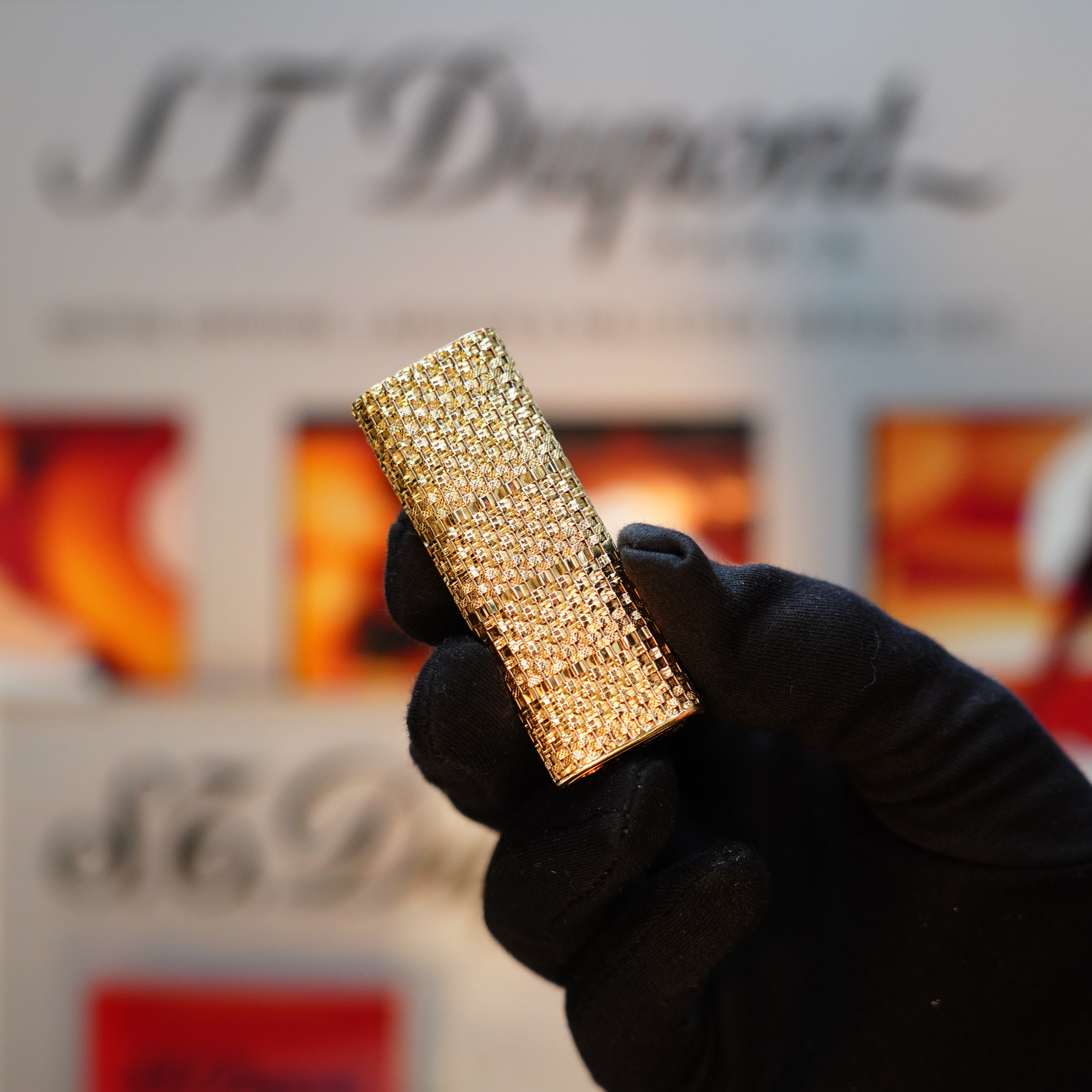 A gloved hand holds the Vintage 1980 Cartier Solid 18k Gold Jacket Basket Lighter, adorned with 750 hallmarks and presented in its display box, against a blurred background of text and colors.