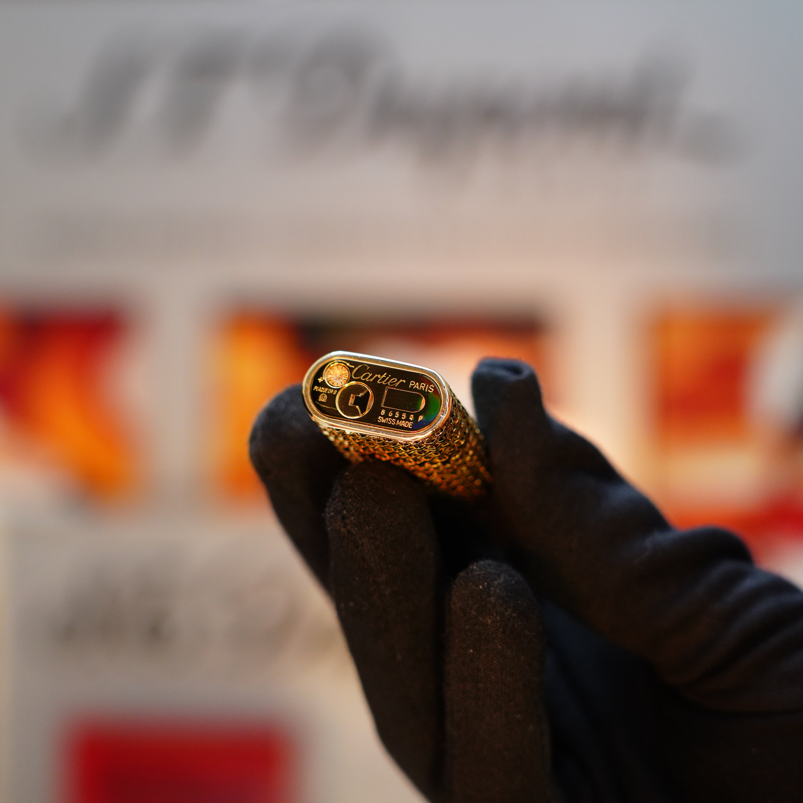 A gloved hand holds a Cartier vintage 1980 Solid 18k Gold Jacket Basket lighter, showcasing the branded roller mechanism. This opulent piece, complete with 750 hallmarks and its display box, emanates luxury against the blurred background.