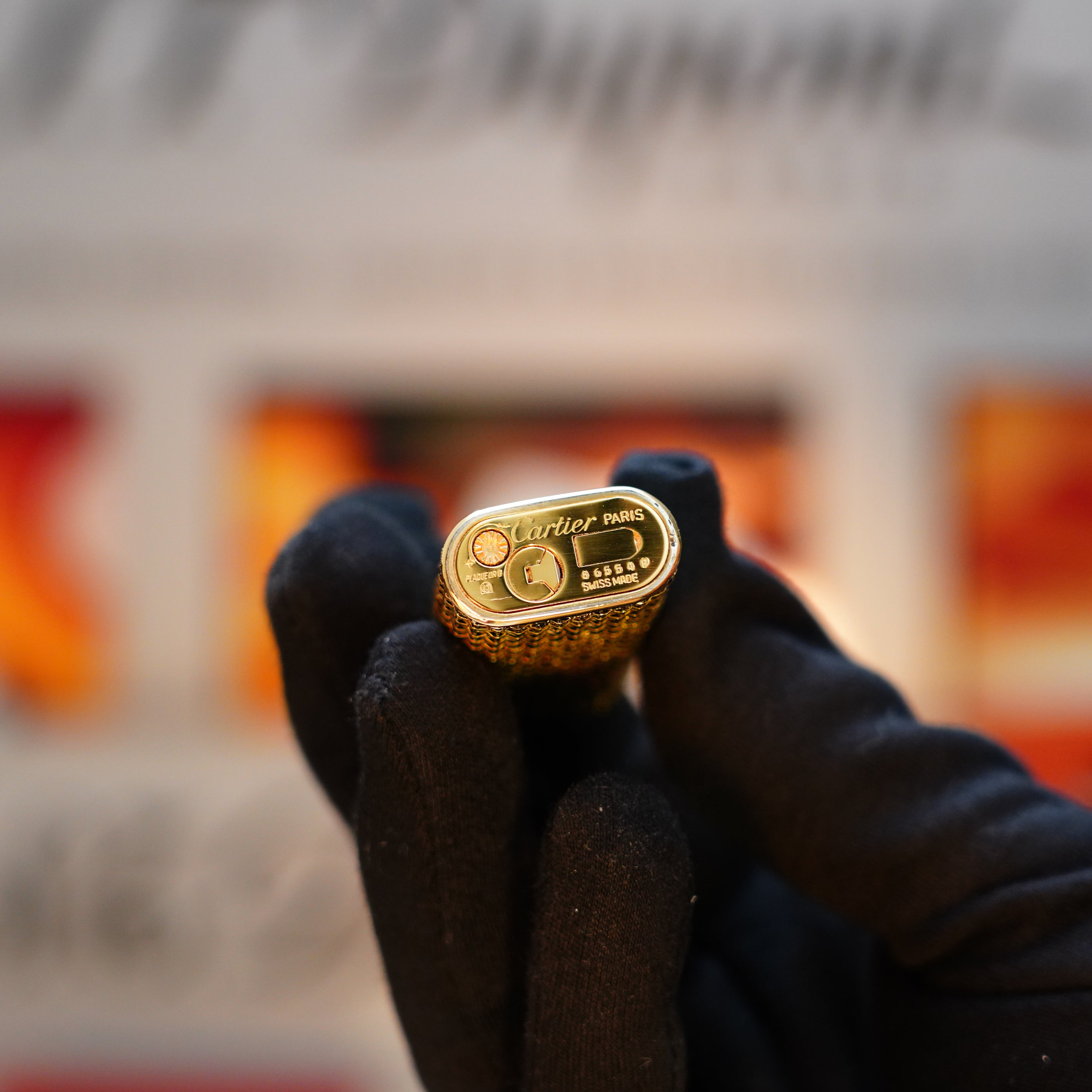 A gloved hand holds a Vintage 1980 Cartier Solid 18k Gold Jacket Basket Lighter adorned with 750 hallmarks, perfectly capturing the elegance of its era. The blurred background enhances the timeless allure of this classic piece, beautifully presented in its display box.