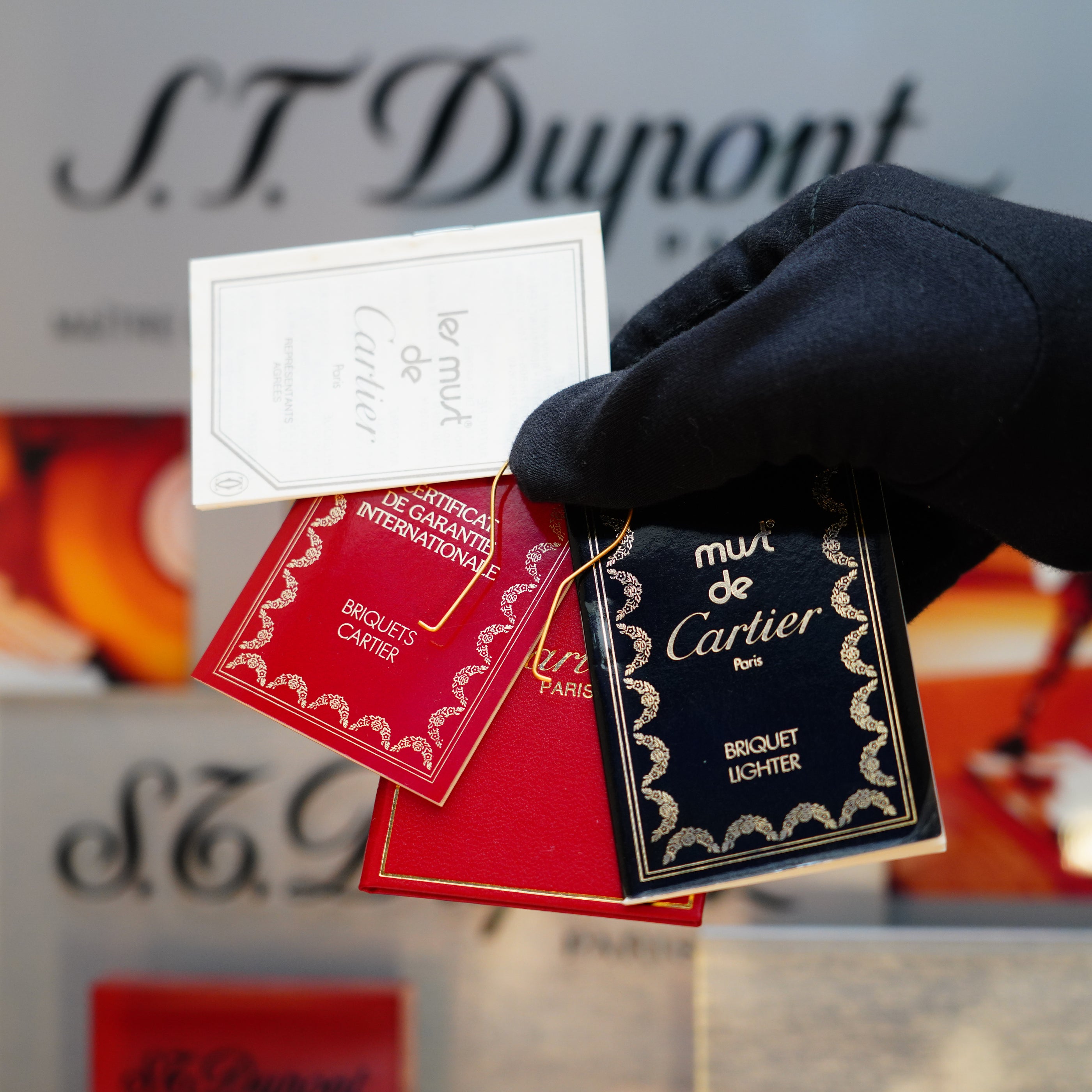 A gloved hand elegantly presents three luxury brand tags against the backdrop of a Vintage 1980 Cartier Solid 18k Gold Jacket Basket Lighter with Display Box, capturing the essence of timeless sophistication.