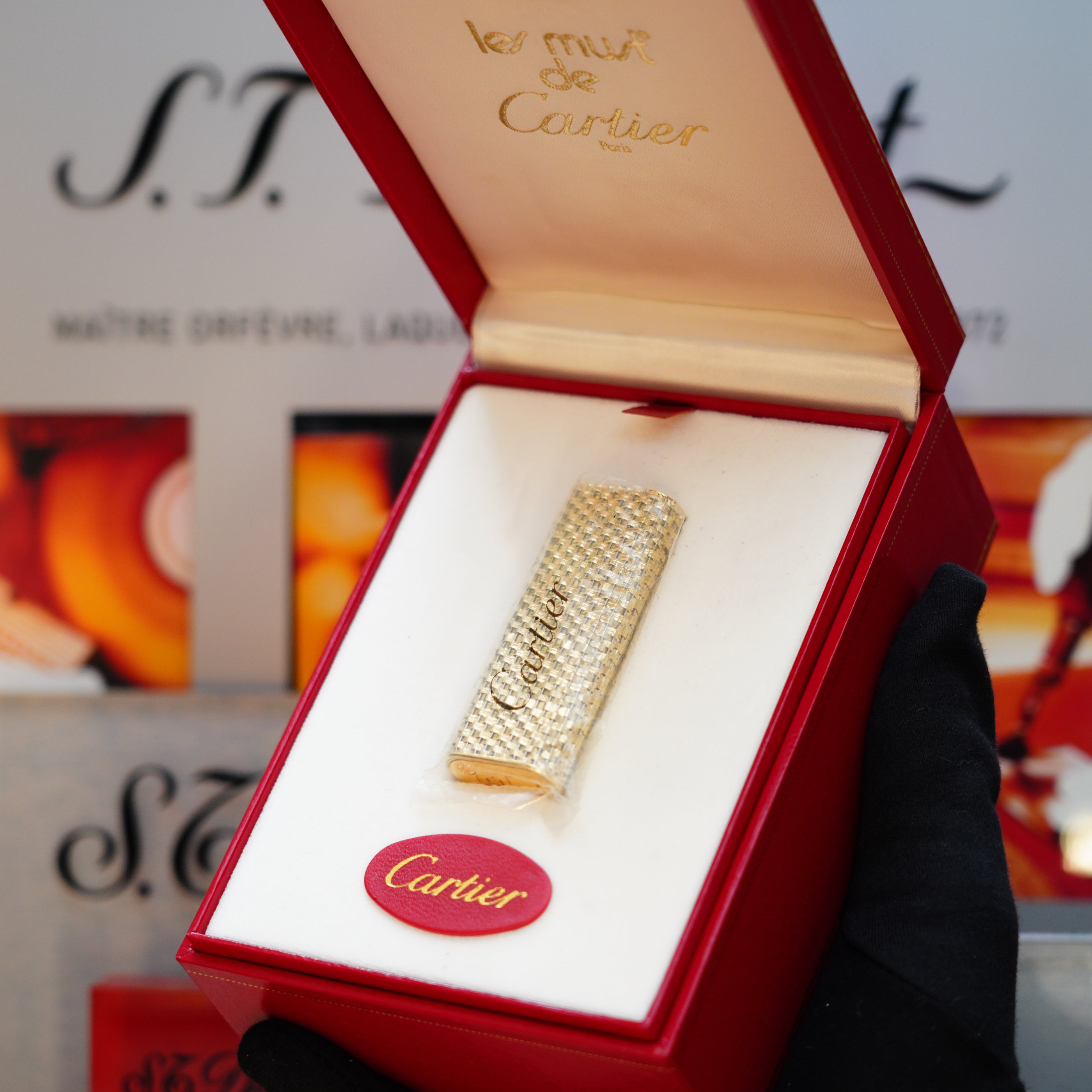 A red Cartier box opens to reveal a vintage 1980 creation by Cartier: a solid 18k gold lighter with 750 hallmarks, intricately encrusted with diamonds and labeled as "les must de Cartier." A gloved hand elegantly showcases this luxurious treasure.