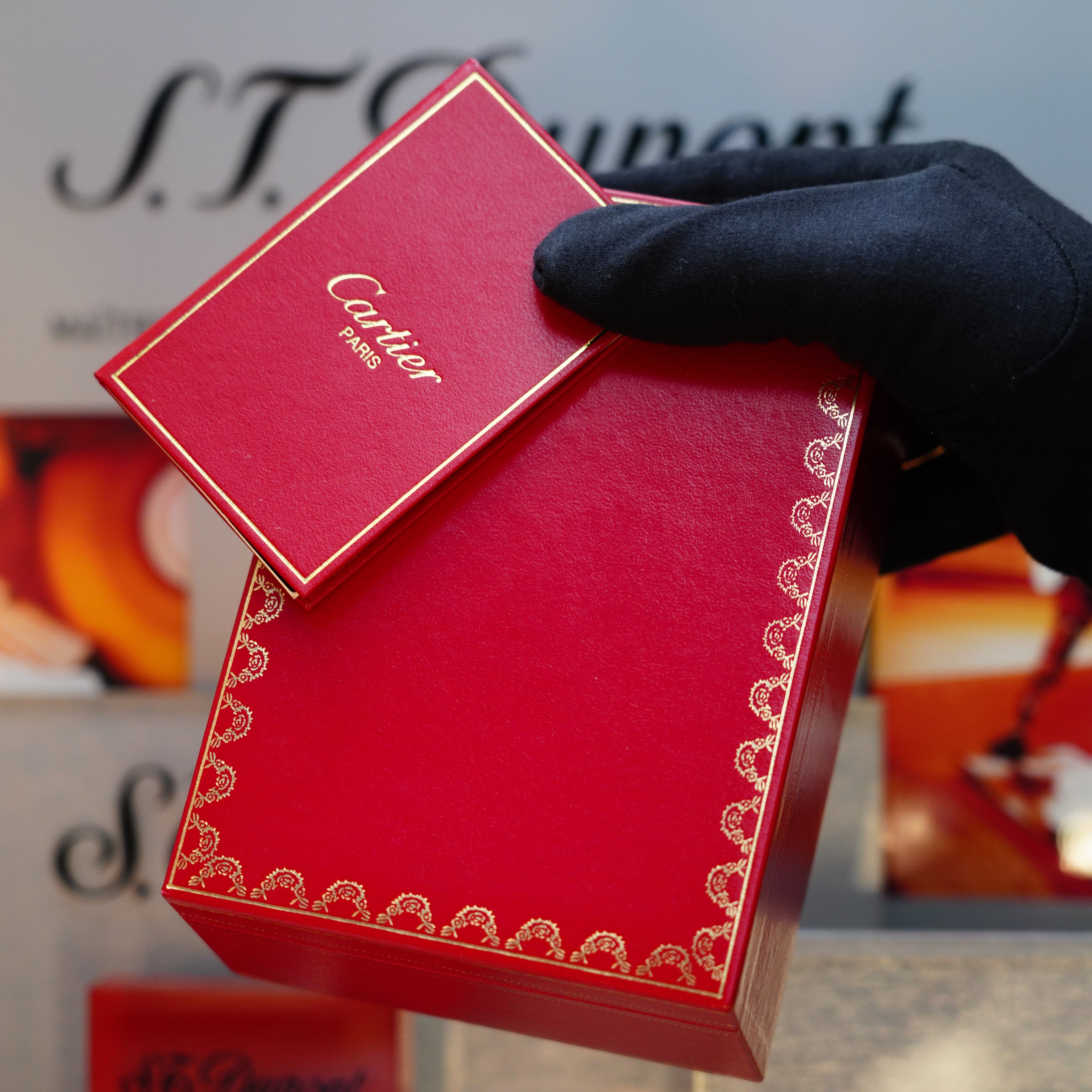 A gloved hand holds a red Cartier box, partially open against a blurred background featuring the S.T. Dupont branding. Inside, there might be a Vintage 1980 Cartier Solid 18k Gold Jacket Basket with 750 hallmarks lighter nestled in the display box.