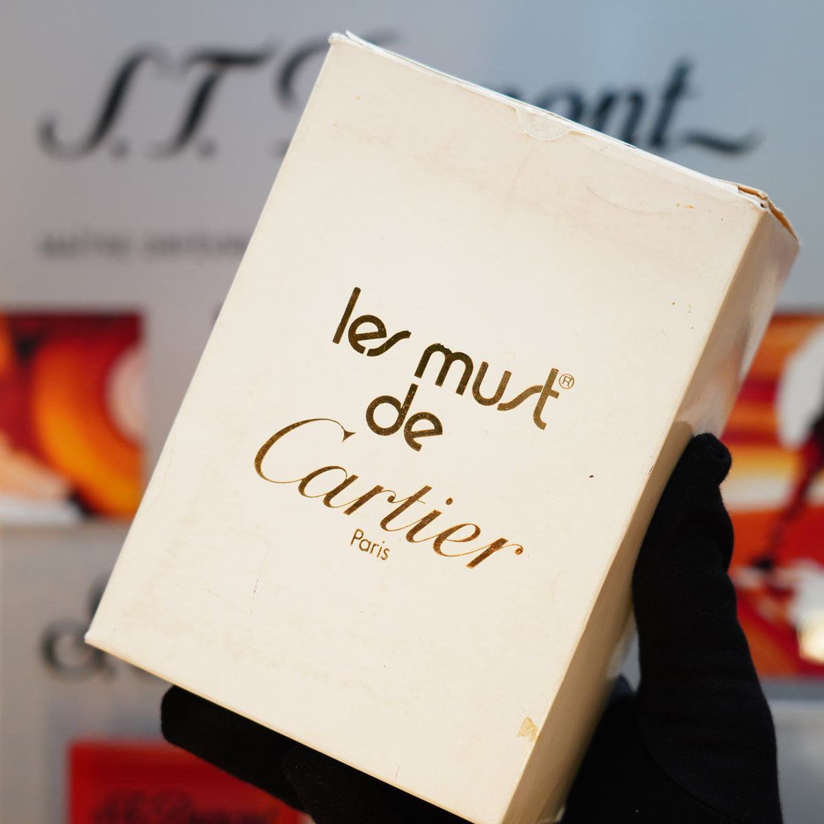 A gloved hand holds a display box labeled with Cartier branding, showcasing the iconic basket design of a vintage 1980 Cartier Solid 18k Gold Jacket Lighter with 750 hallmarks, set against a blurred background with signage.