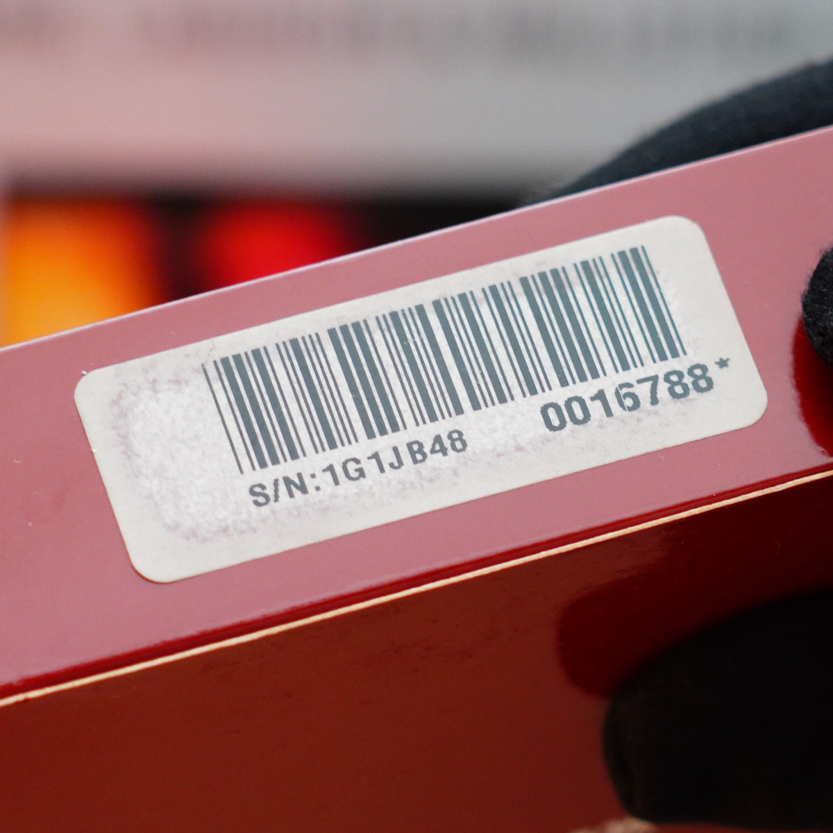 A gloved hand holds a red object with a white label, displaying a barcode and text "S/N:1G1JB48" and "0016788*". Its sleek design is reminiscent of the Vintage 1993 S.T. Dupont Noire Heavy Palladium Finish Placage, Bandes De LAQ Ligne 2 Lighter Double Flame, echoing the elegance of S.T. Dupont's craftsmanship.