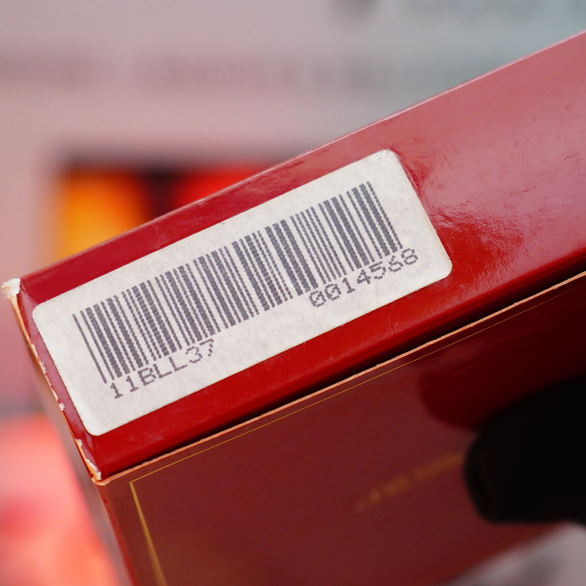 A close-up of a red box with a barcode sticker showing the numbers "0014558 11BLL37," its vibrant color echoing that of the Vintage 1990 S.T. Dupont Rare Contemporaine Ligne 1 Green Vert Filet Plaque Lacquer lighter. The blurred background enhances the scene's mysterious allure.