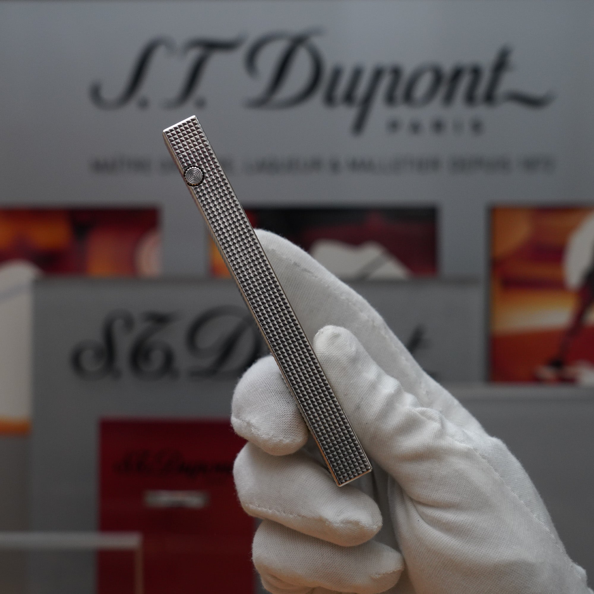 A vintage hand holds a Dunhill Silver plated piece of metal in front of an st dupont sign.