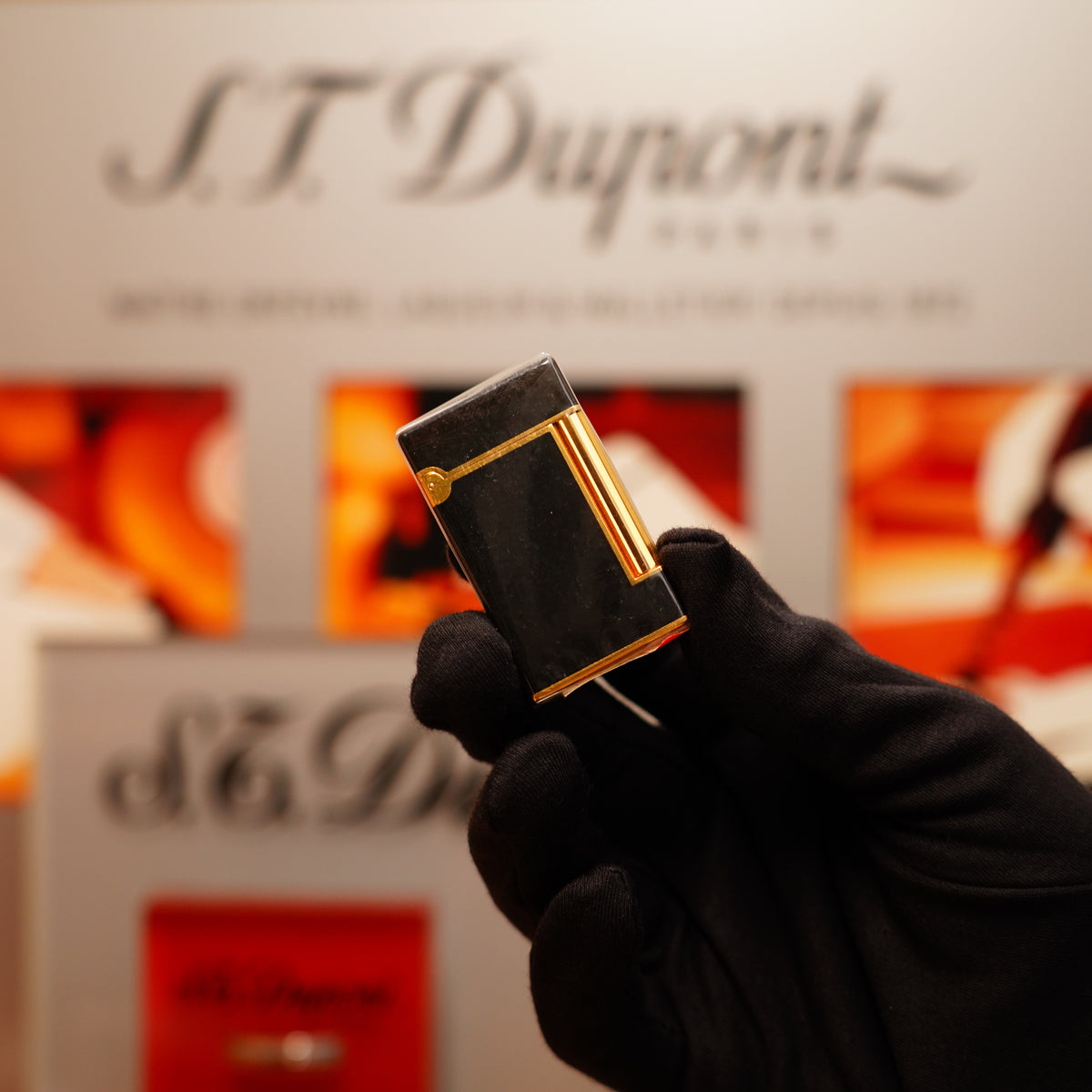 A gloved hand holds a Vintage 1990 S.T. Dupont Overall Black Lacquer Early 24k Gold Ligne D Sealed Lighter against a blurred backdrop of similar luxury items.