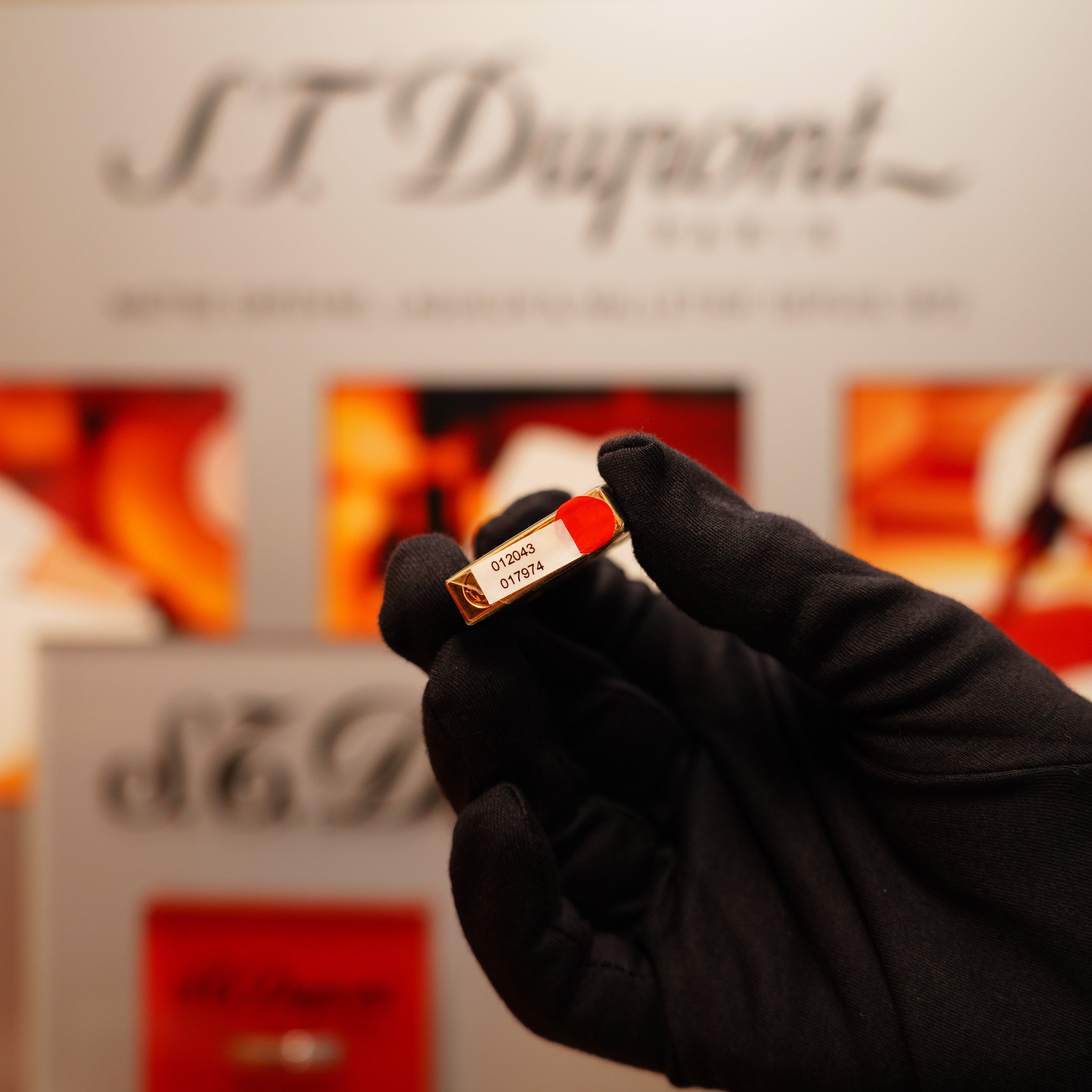 A gloved hand holds a red lipstick in the foreground, with the blurred branding of S.T. Dupont's Vintage 1990 Overall Black Lacquer Early 24k Gold Ligne D Sealed Lighter in the background.