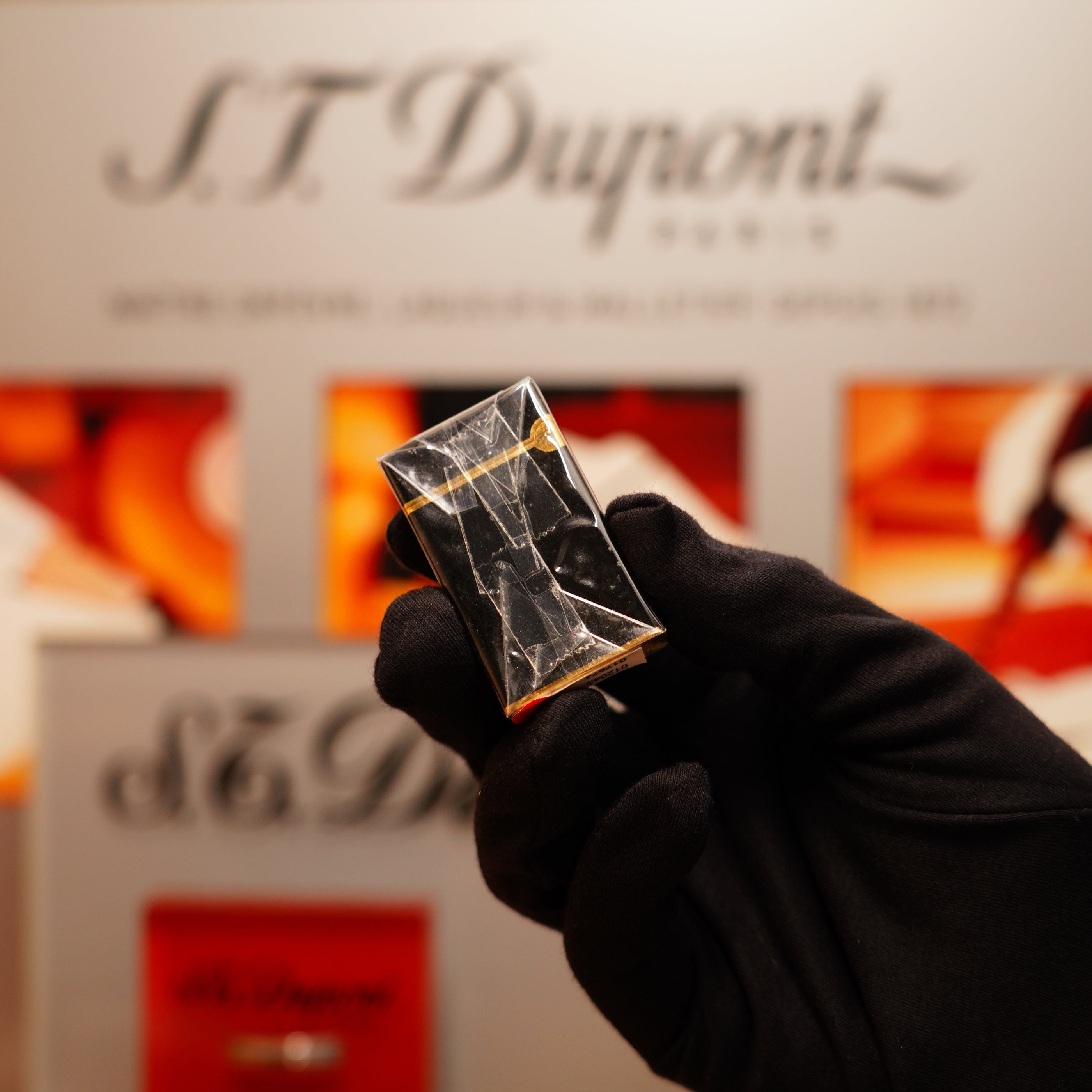 A gloved hand holds a small, transparent, rectangular object identified as a Vintage 1990 S.T. Dupont Overall Black Lacquer Early 24k Gold Ligne D Sealed Lighter with a blurred branded background.
