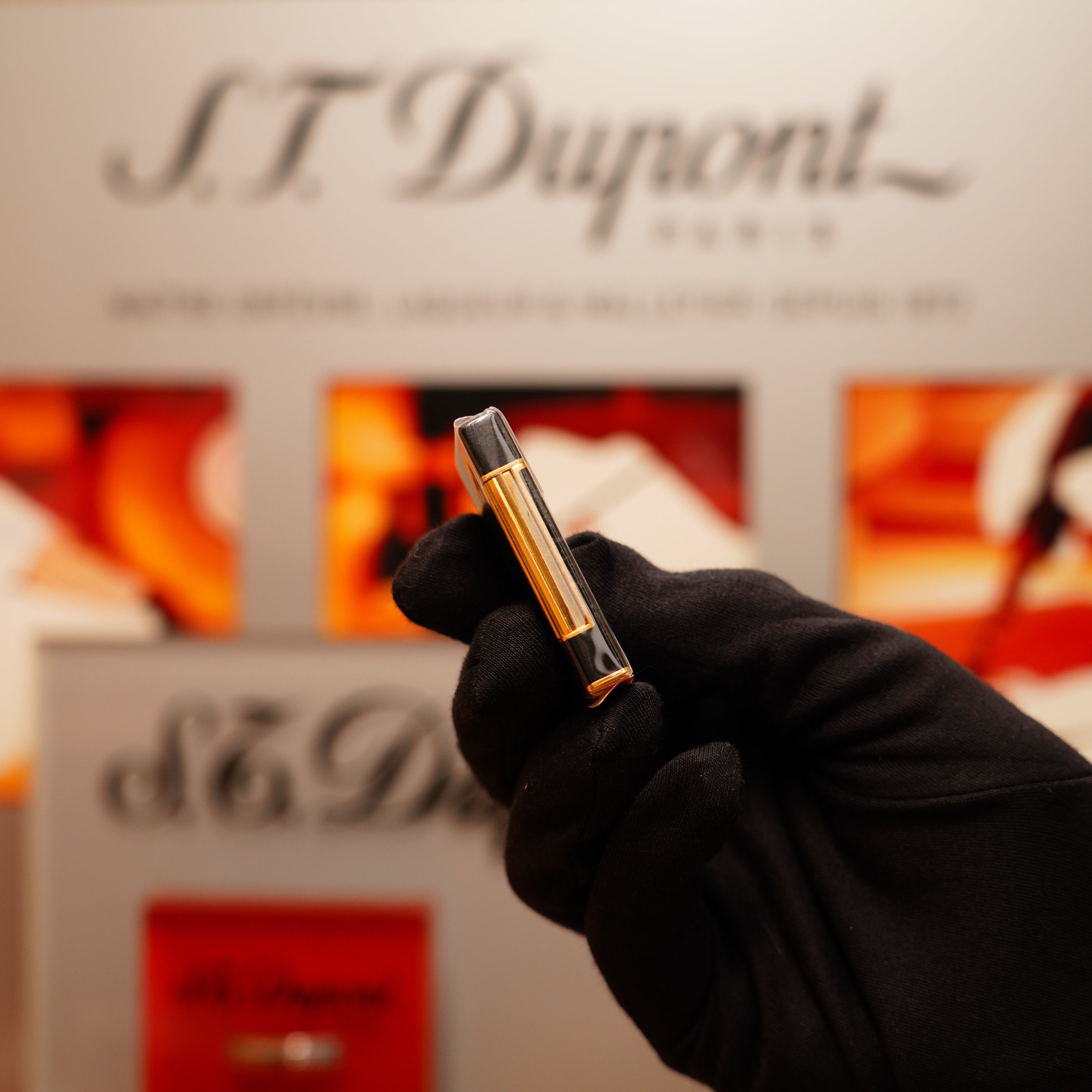 A gloved hand holds a small, black lacquer and 24k gold lighter with an S.T. Dupont backdrop in the background.