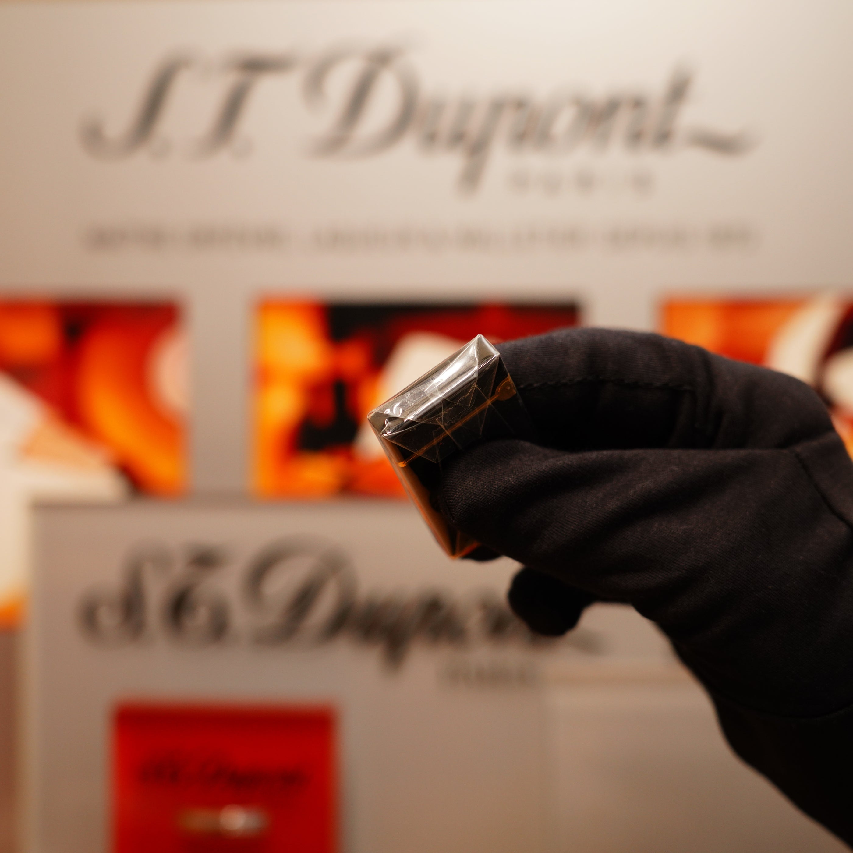 A gloved hand holds a Vintage 1990 S.T. Dupont Overall Black Lacquer Early 24k Gold Ligne D Sealed Lighter against a blurred background featuring the S.T. Dupont logo.