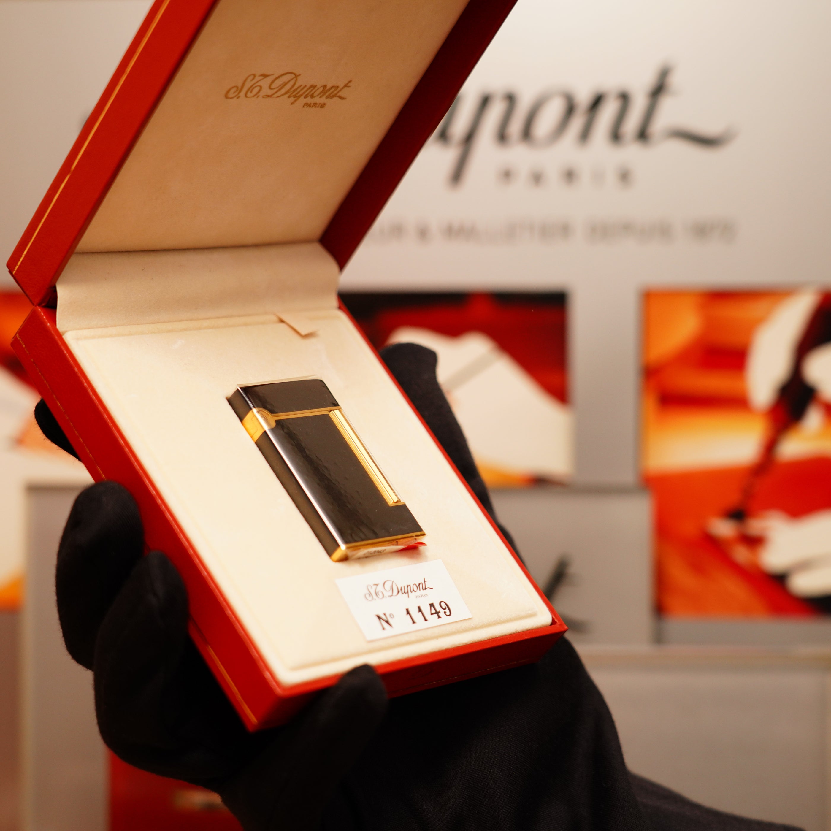 A person wearing black gloves holds an open red box showing a black and gold lighter. The box lid reads "S.T. Dupont Paris," and the lighter is labeled "N° 1149." It's a Vintage 1990 S.T. Dupont Overall Black Lacquer Early 24k Gold Ligne D Sealed Lighter.