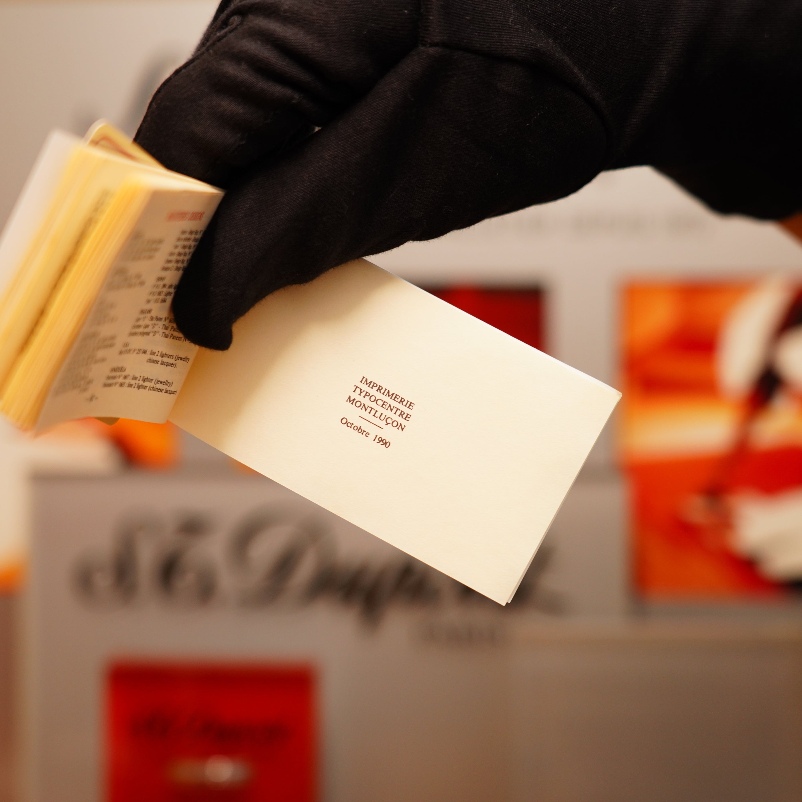 A gloved hand holds an open small book with visible text, alongside a Vintage 1990 S.T. Dupont Overall Black Lacquer Early 24k Gold Ligne D Sealed Lighter.
